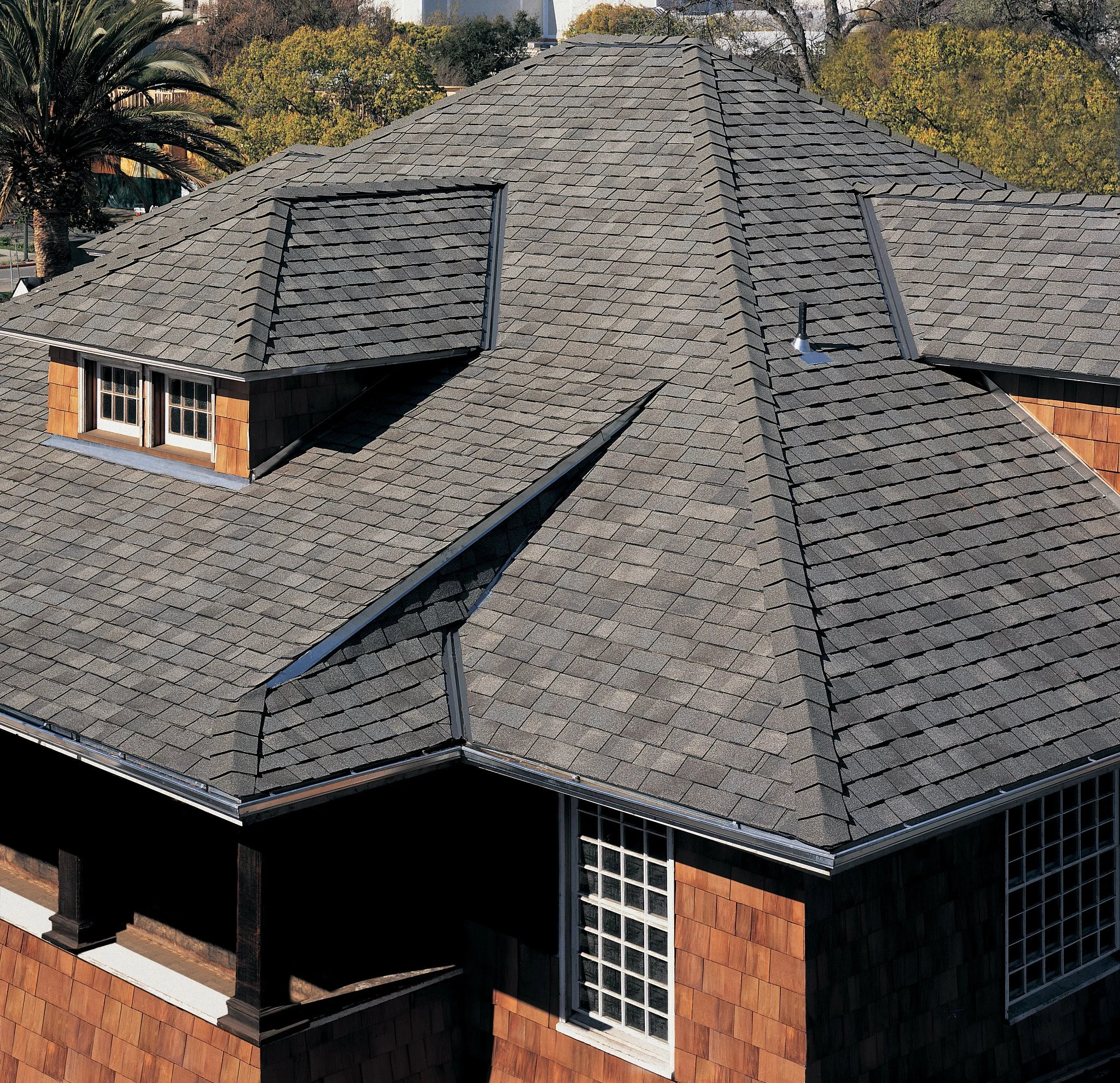 Aerial view of a house with gray shingle roof and small dormer window surrounded by trees with autumn foliage.