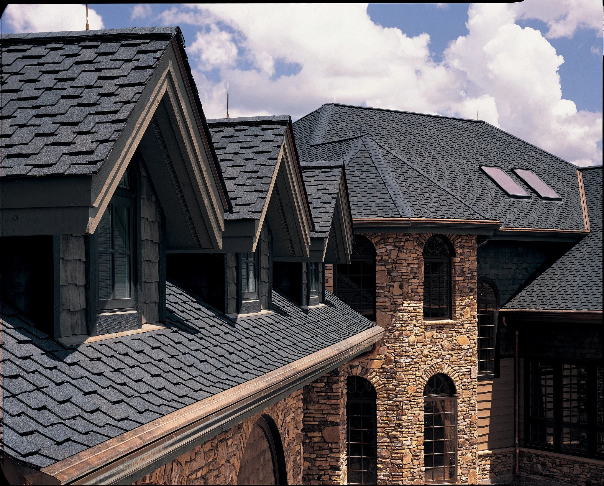 Close-up of gray shingle roof tiles on multiple houses under a partly cloudy sky.