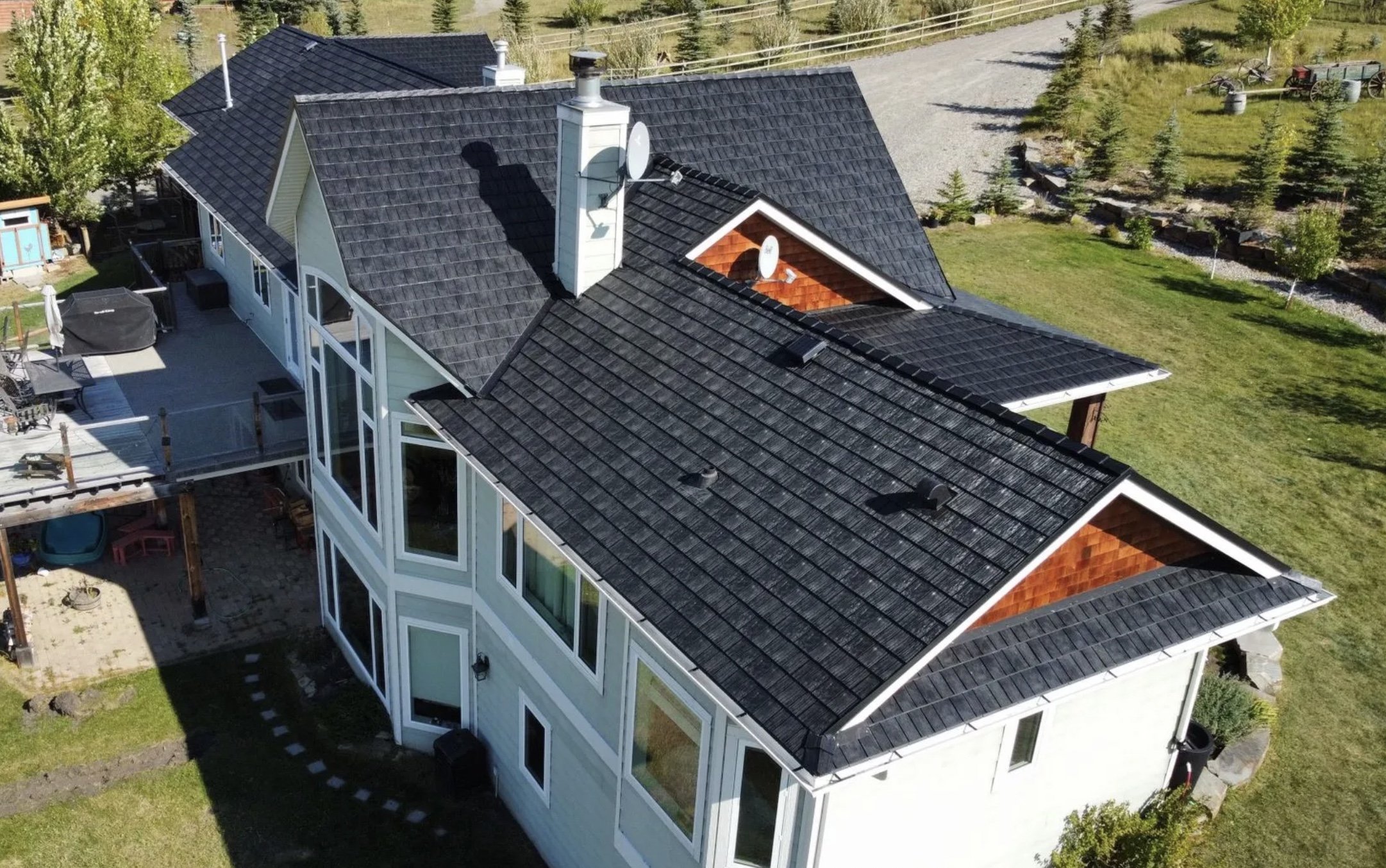 Aerial view of a modern house with a large gray shingled roof, multiple chimneys, and satellite dishes, surrounded by a green yard with trees, a gravel pathway, and outdoor furniture.
