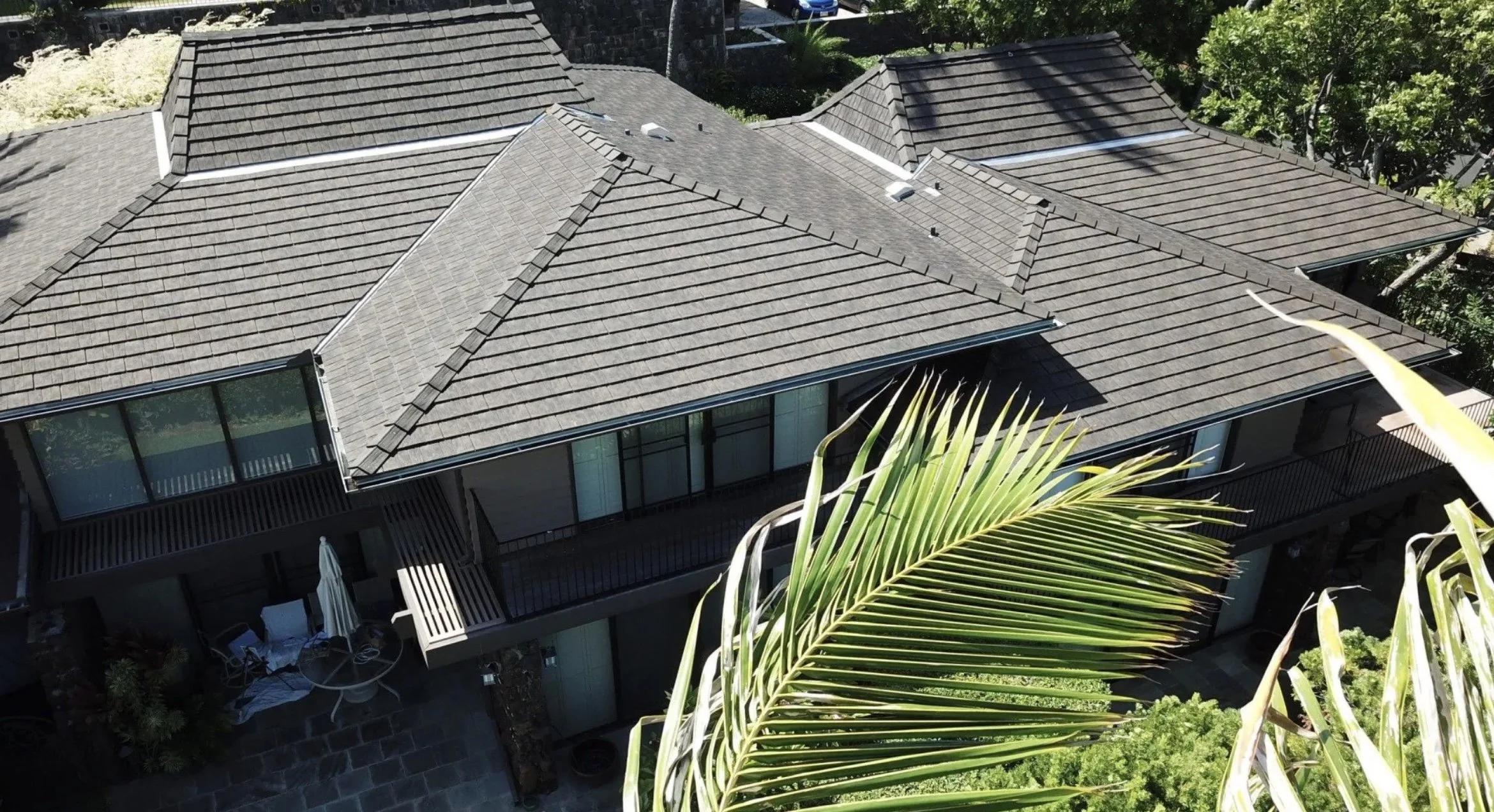 Aerial view of a multi-story house with a gray sloped roof and large windows, surrounded by trees and greenery.