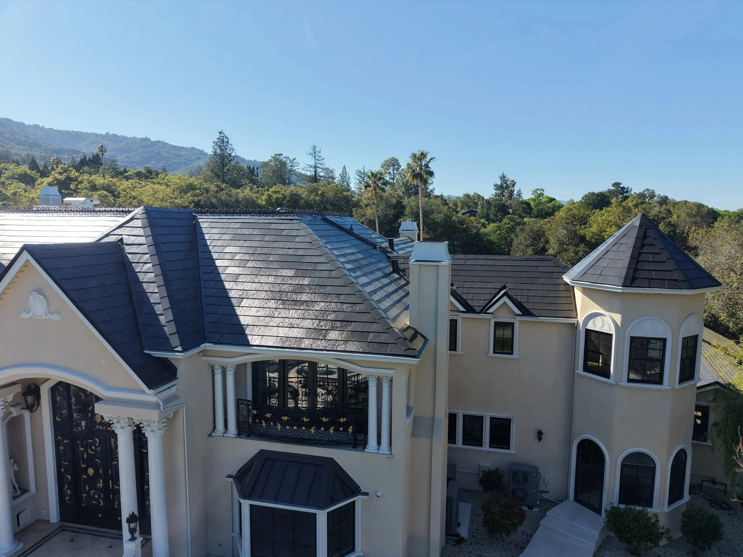 A large, elegant house with dark gray tiled roofs, white walls, and decorative black wrought-iron balconies, set against lush green trees and distant mountains under a clear blue sky.