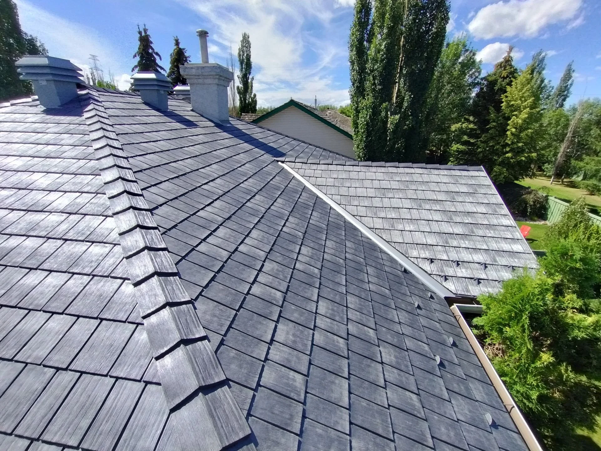 View of a house roof with wooden shingles, surrounded by tall trees and a blue sky with some clouds.