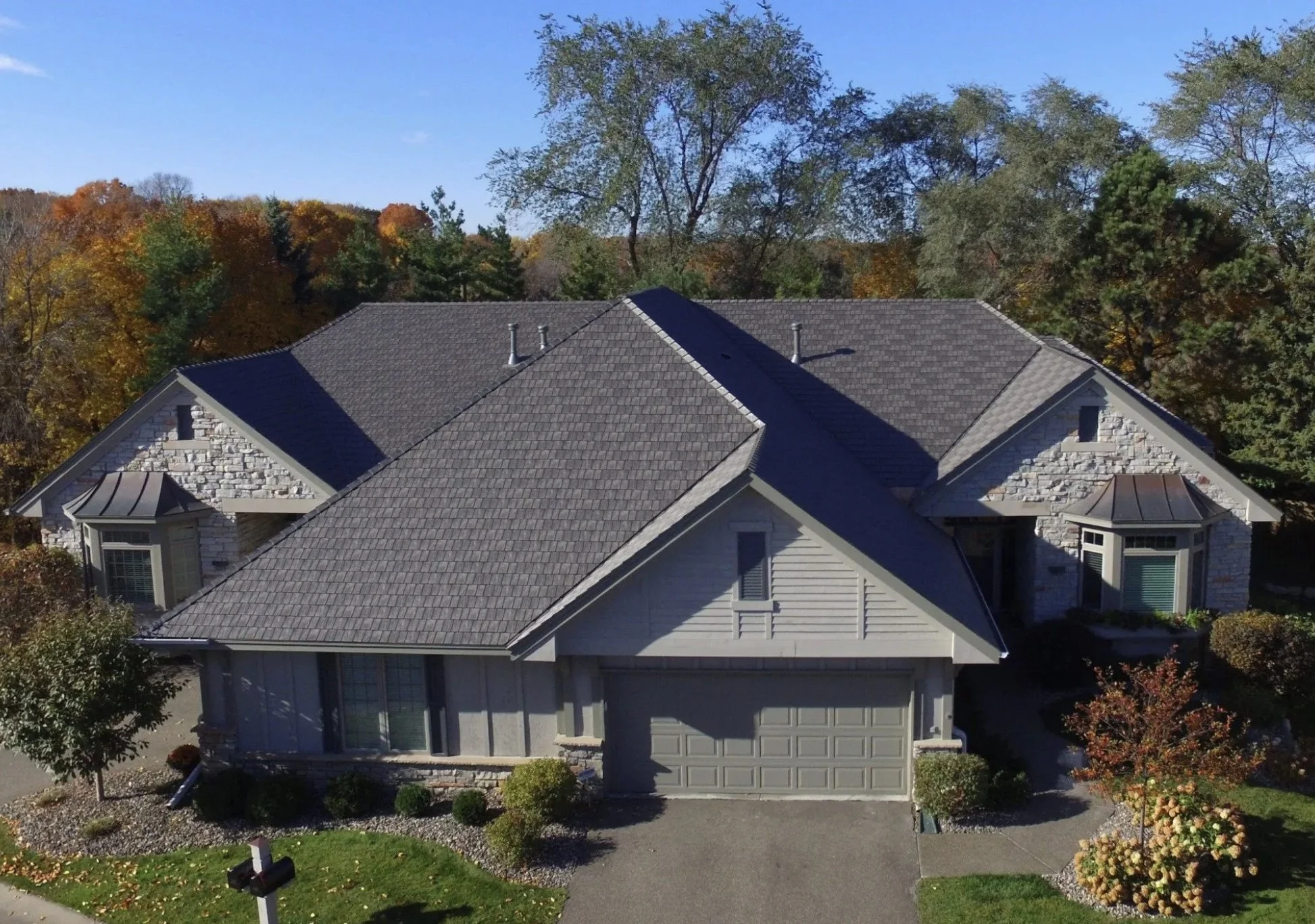 A house with a grey shingle roof and white exterior, surrounded by trees with fall foliage, under a blue sky.