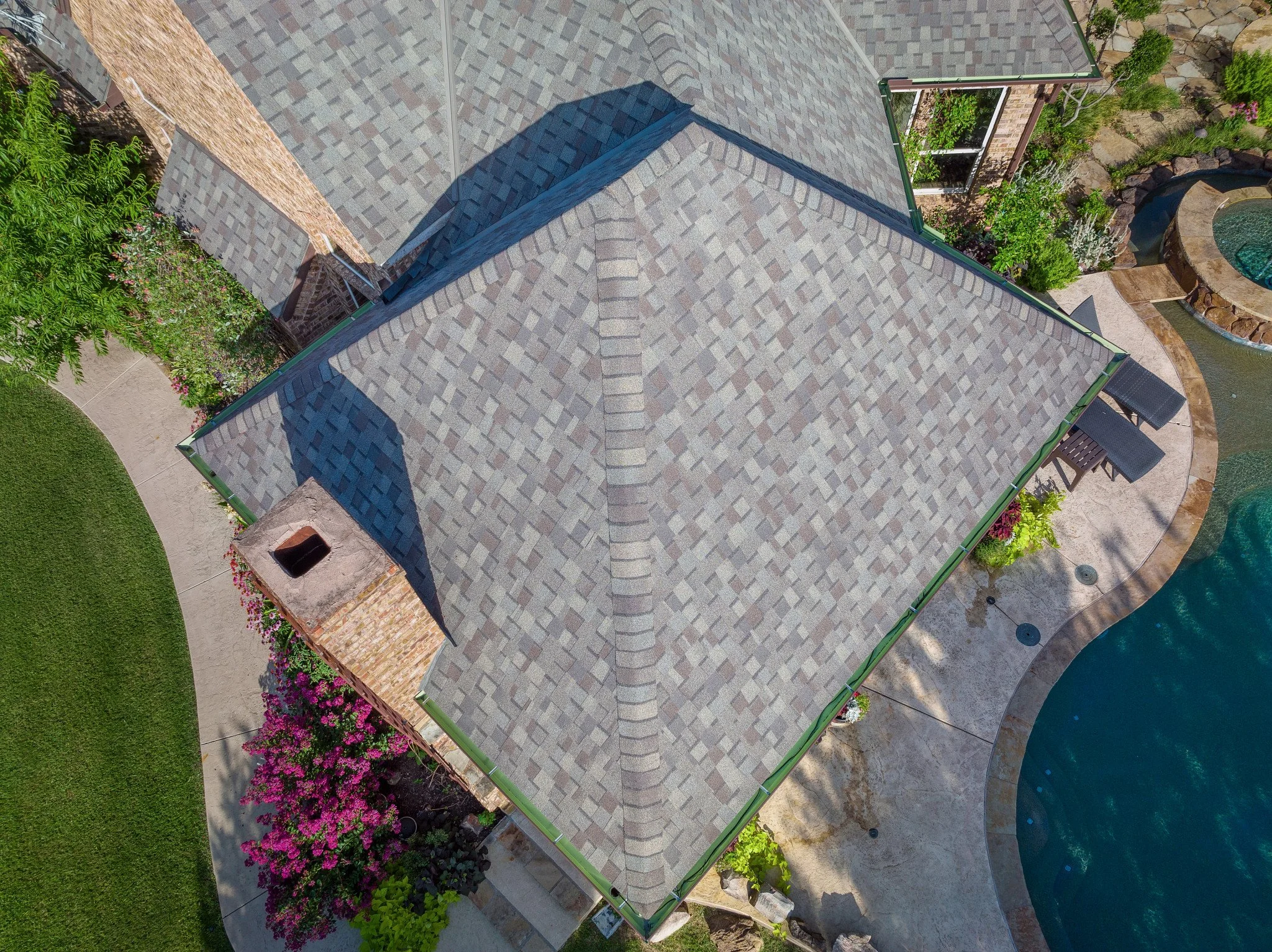 An aerial view of a house with a gray shingled roof, surrounded by a landscaped garden with pink flowers, green plants, a small pond, and a patio area with outdoor furniture.