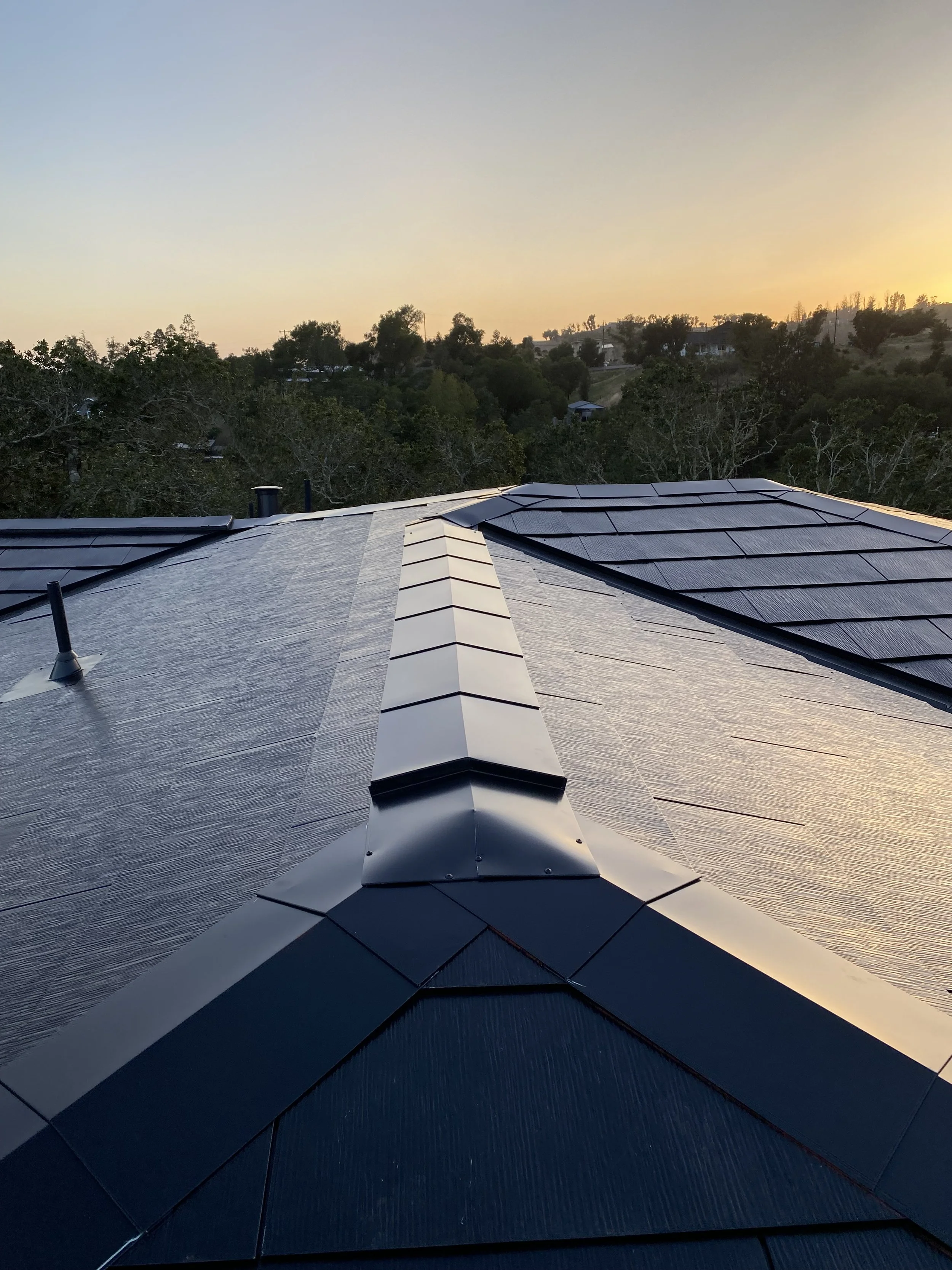 Metal roof with black shingles and a ridge vent, with trees and houses in the background at sunset.