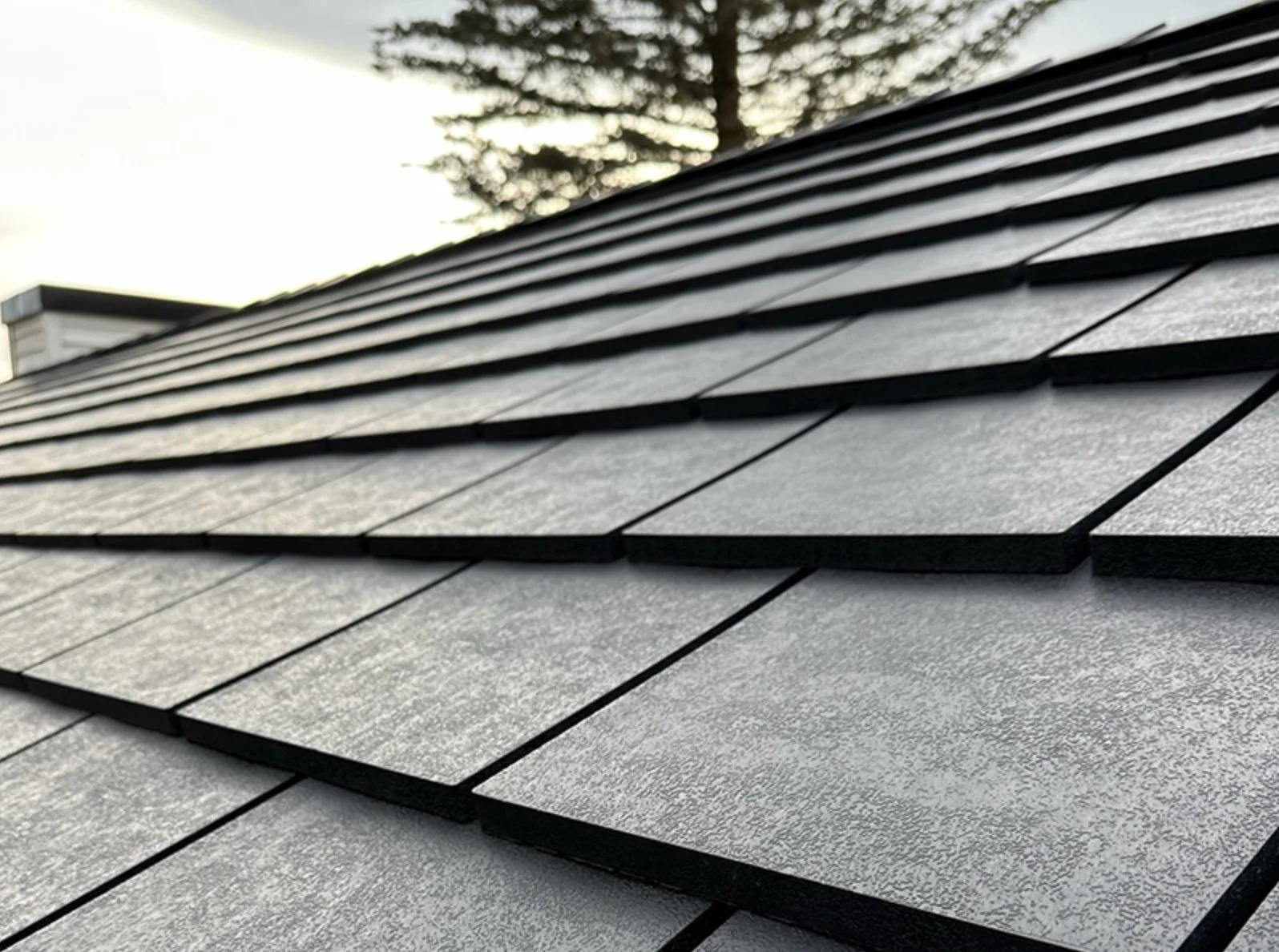 Close-up of a sloped roof with gray asphalt shingles and black trim, with a tree and cloudy sky in the background.