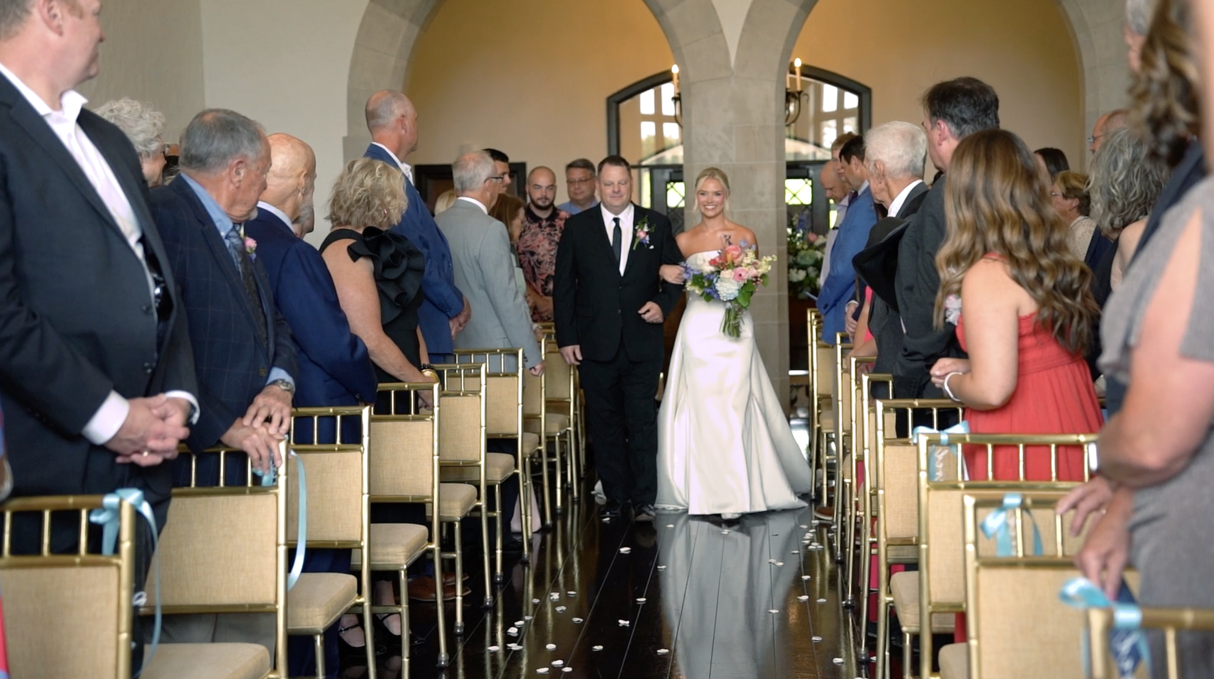 A bride in a white wedding dress holding a bouquet walks down the aisle with a man in a black suit. Guests are seated on both sides, watching her walk. The setting is an indoor wedding ceremony with large arched windows in the background.