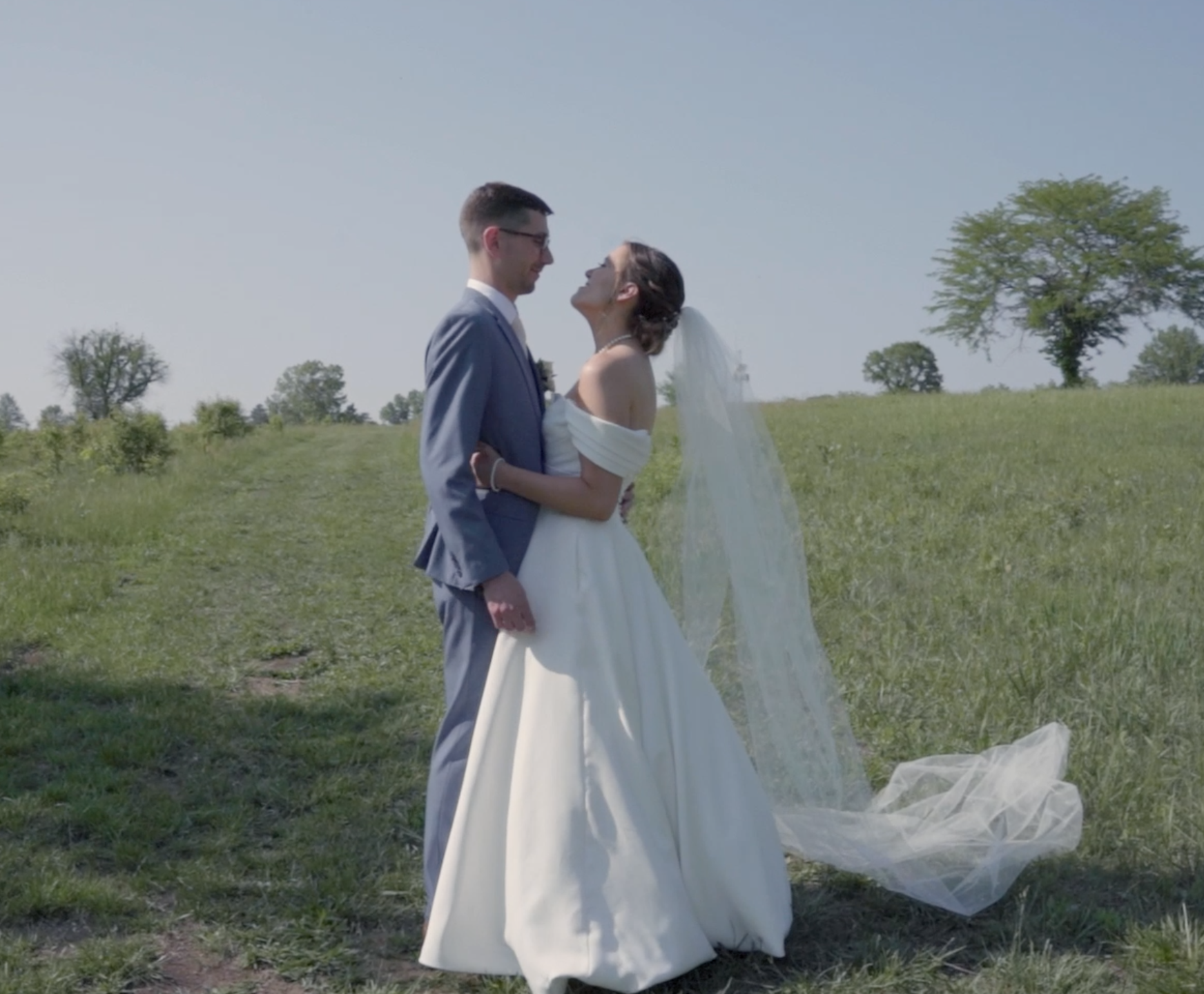 A bride and groom standing in a grassy field, embracing and smiling at each other on their wedding day, with trees and a clear blue sky in the background.