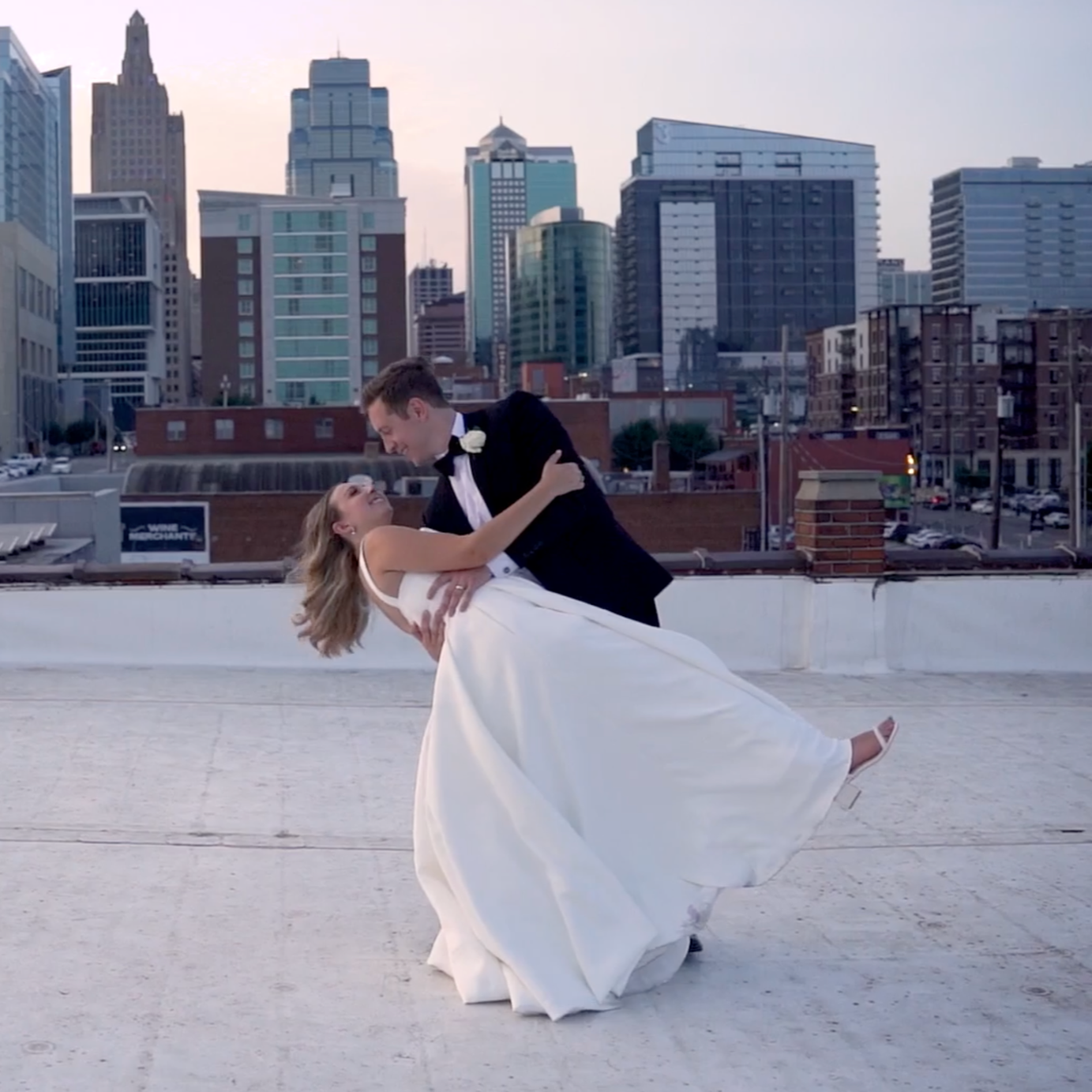 A bride and groom dance on a rooftop with a city skyline in the background, during sunset.