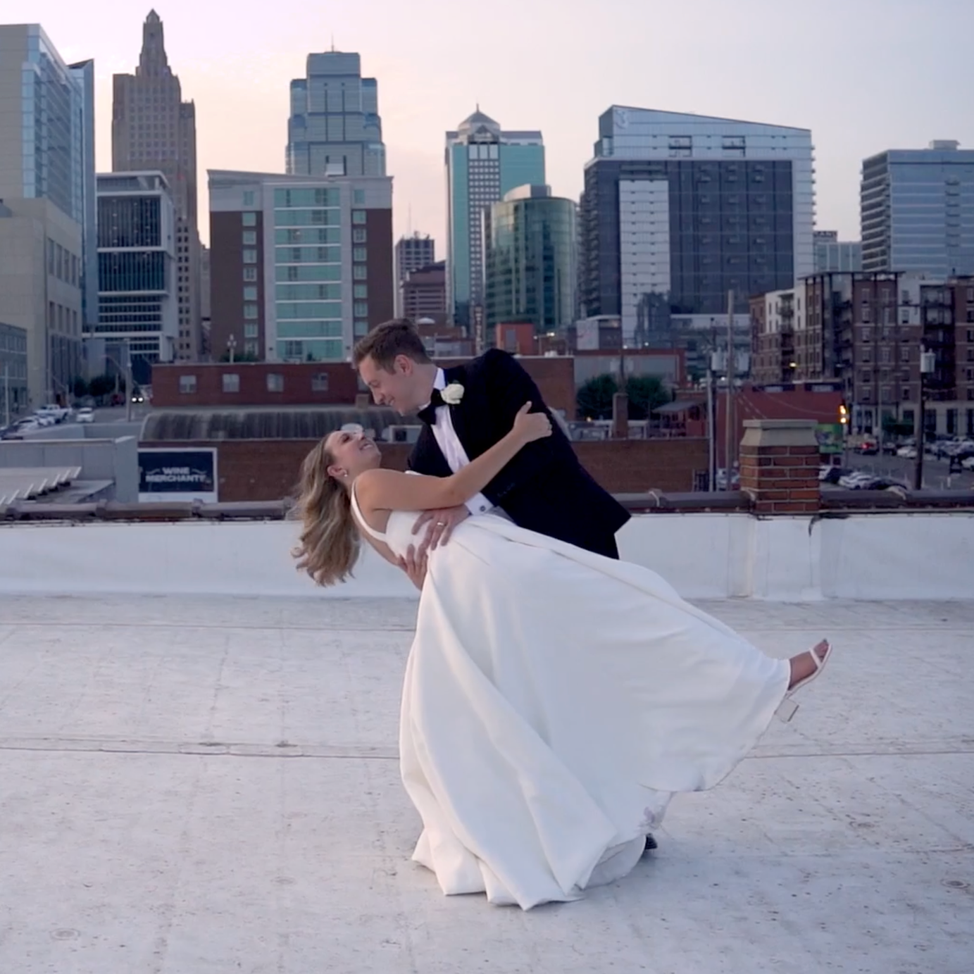 A couple dancing on a rooftop with a city skyline in the background during sunset, the man in a tuxedo and the woman in a white wedding dress.