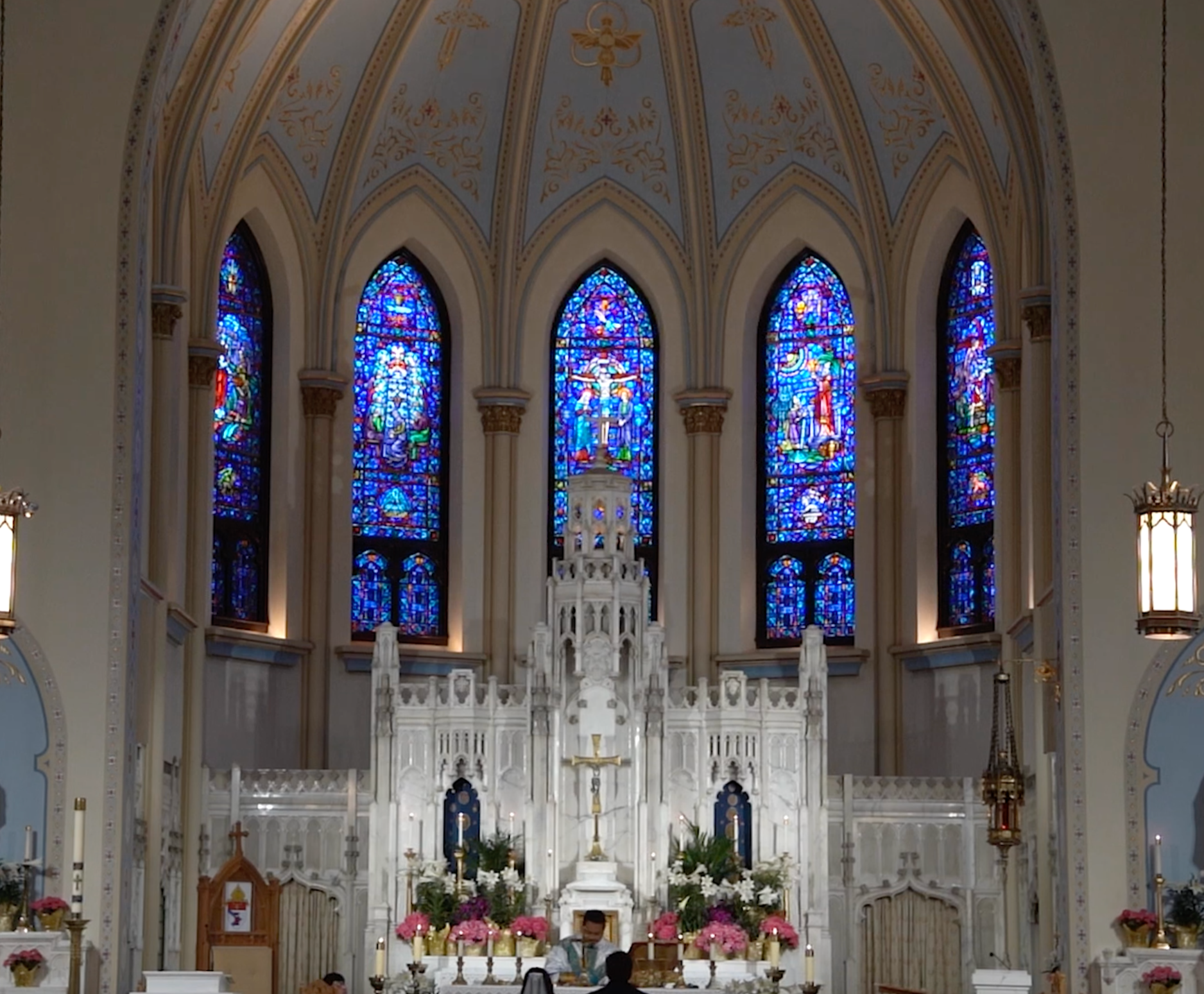 Interior of a church with stained glass windows, an altar with a crucifix, and colorful flowers, with a person standing at the altar.