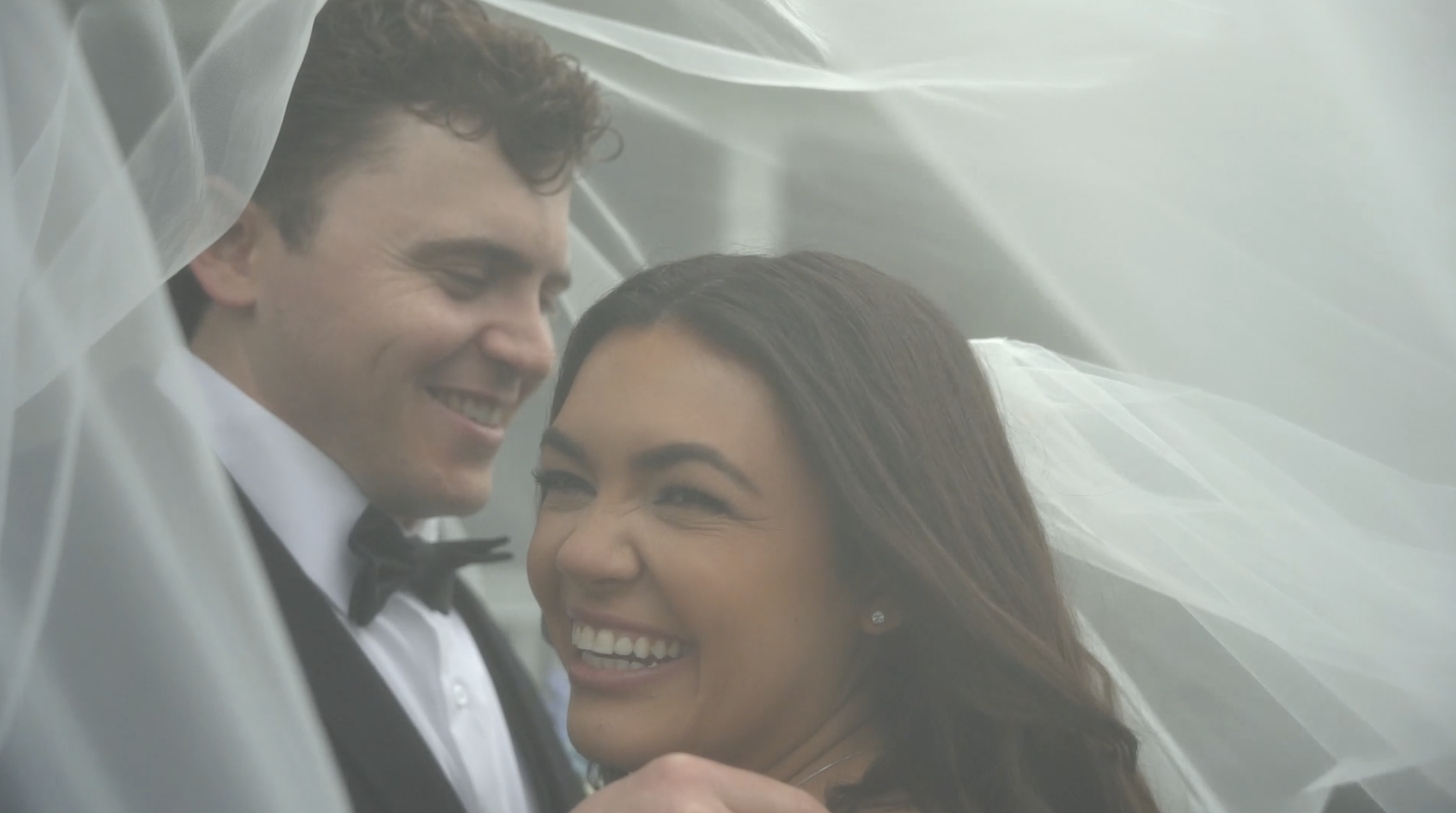 A smiling couple, dressed in wedding attire, sharing an intimate moment under a veil.