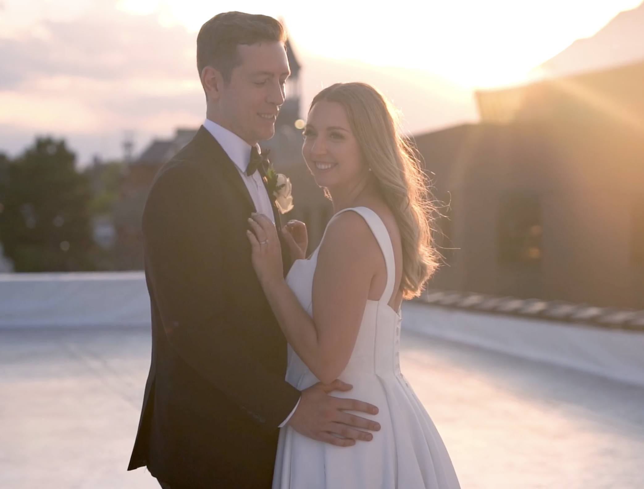 A happy couple in wedding attire stands close together on a rooftop at sunset, with the man in a tuxedo and the woman in a white wedding dress, smiling at each other.