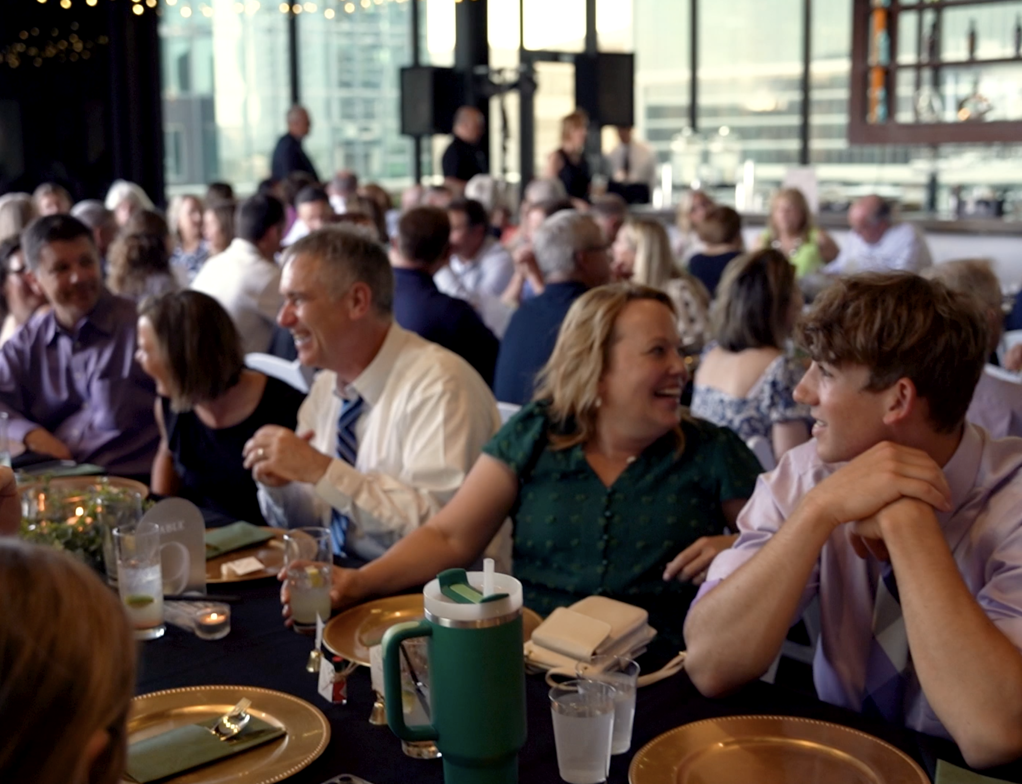People sitting and talking at a banquet table during a social event or gathering in a restaurant or banquet hall with large windows and city buildings in the background.