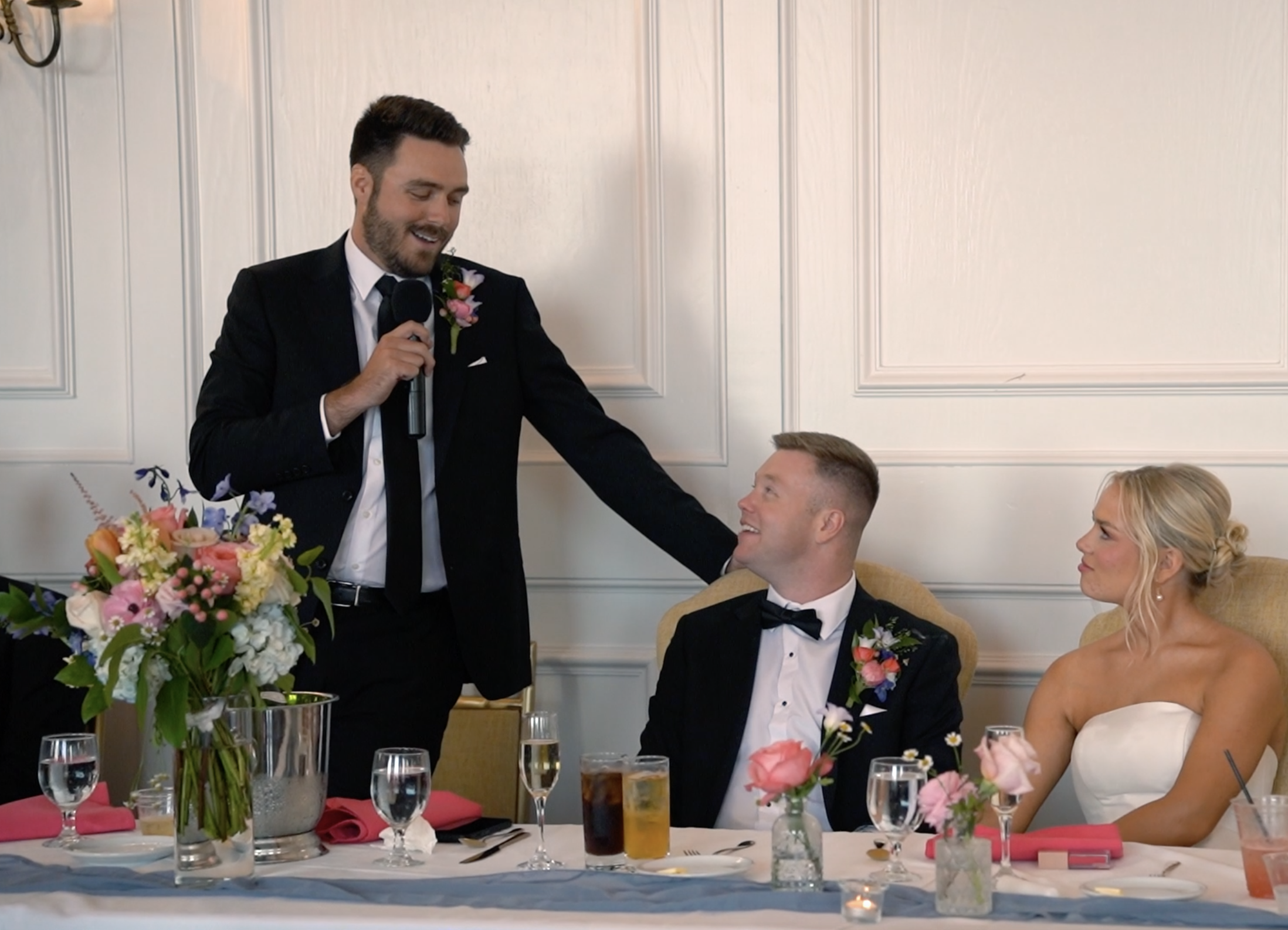 At a wedding reception, a man in a tuxedo is giving a speech, standing next to a seated groom and bride. The groom is wearing a tuxedo with a bow tie, and the bride is in a strapless wedding dress. There are flowers and drinks on the table.
