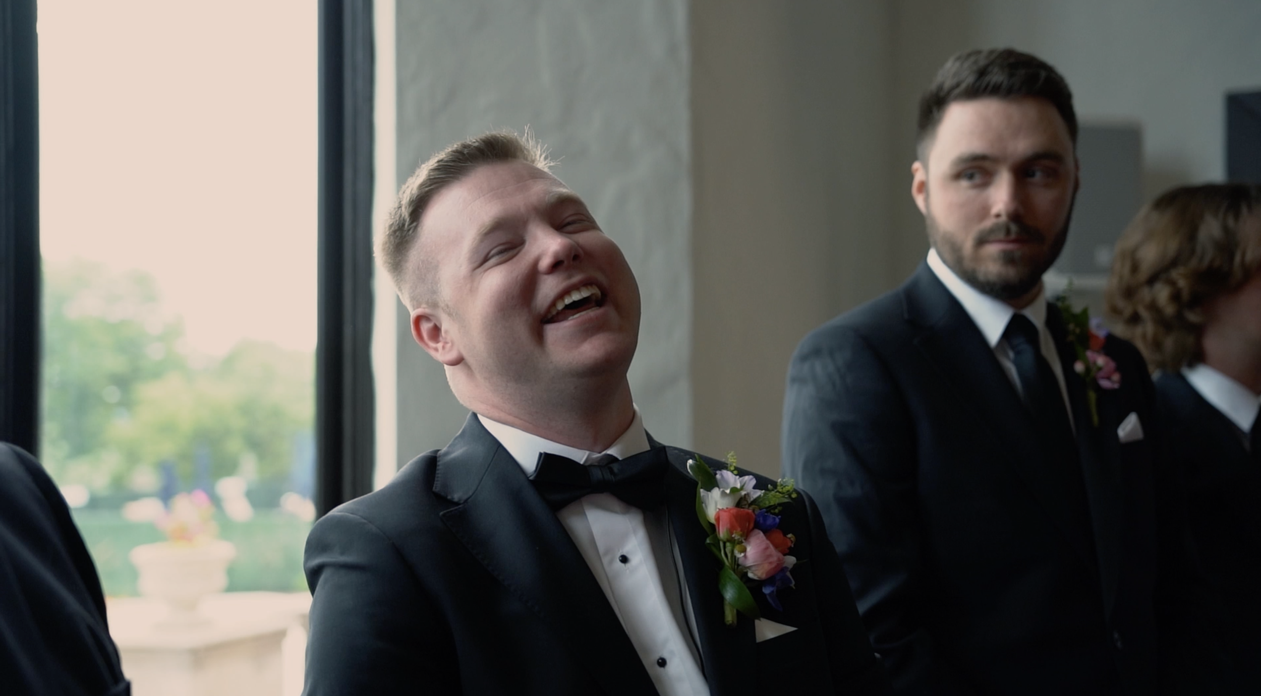 A man in a tuxedo and bow tie is smiling and laughing during a wedding ceremony, standing next to another person in formal attire.