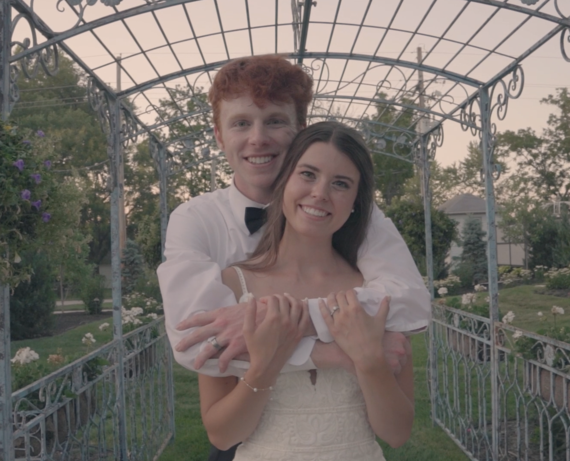 A happy couple dressed in wedding attire, outdoors in a garden with purple flowers and trees, posing under a metal arbor at sunset.