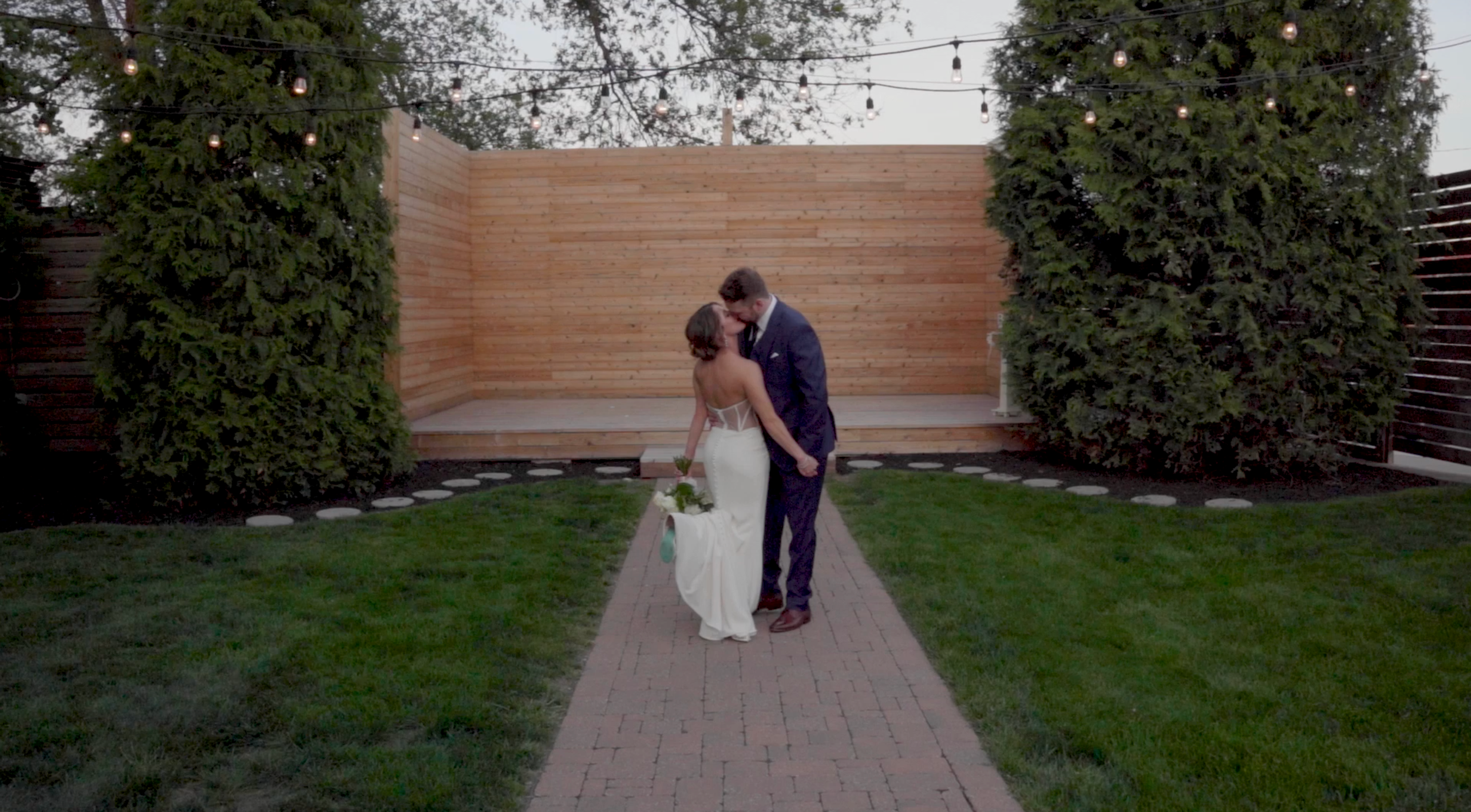 A bride and groom sharing a kiss on a brick path in a backyard, with string lights overhead, tall trees on each side, and a wooden fence and wall in the background.