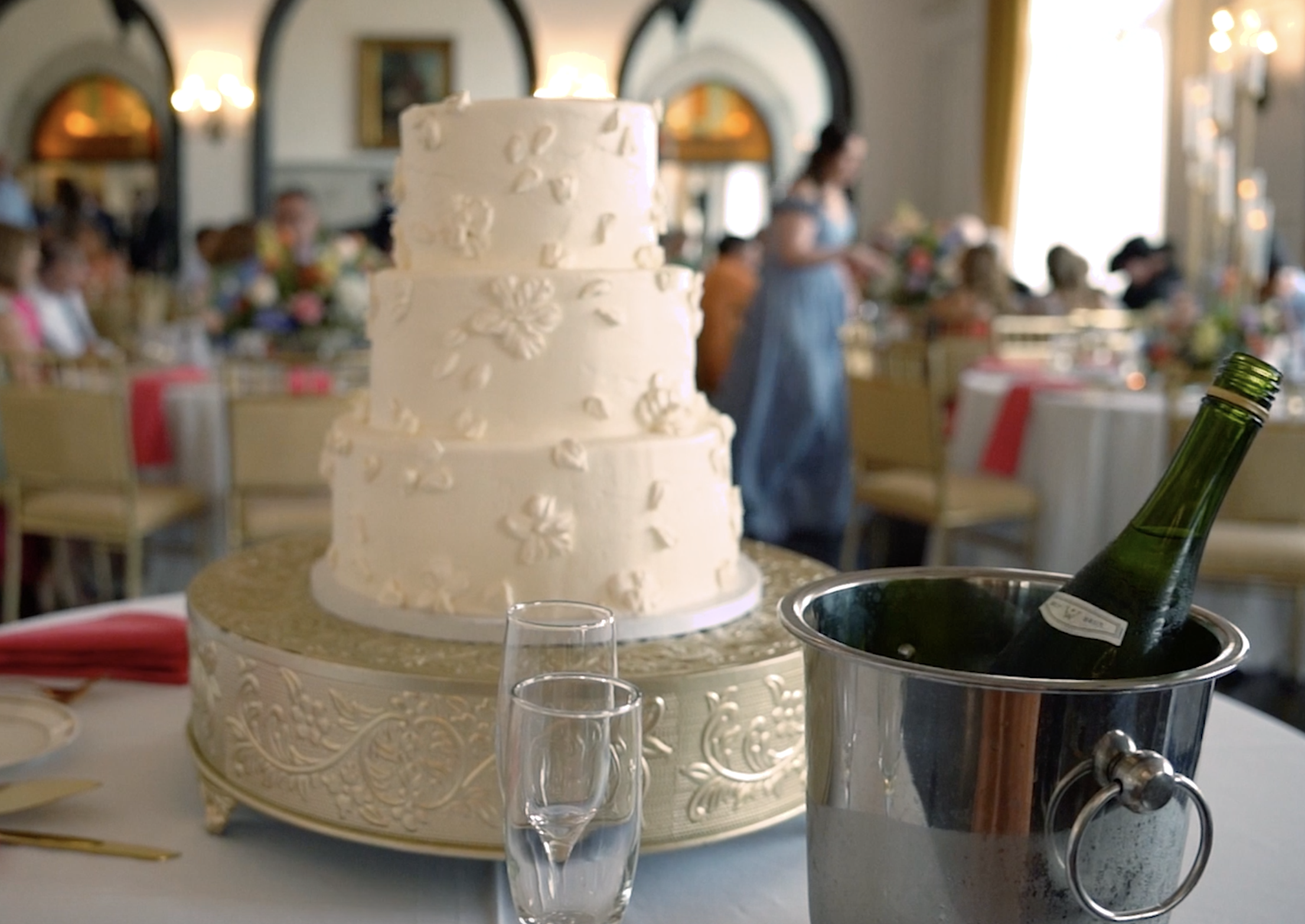 A three-tier white wedding cake with floral decorations on a decorated cake stand, a champagne bottle in an ice bucket, and two champagne glasses on a wedding reception table, with blurred guests and a woman in a blue dress in the background.