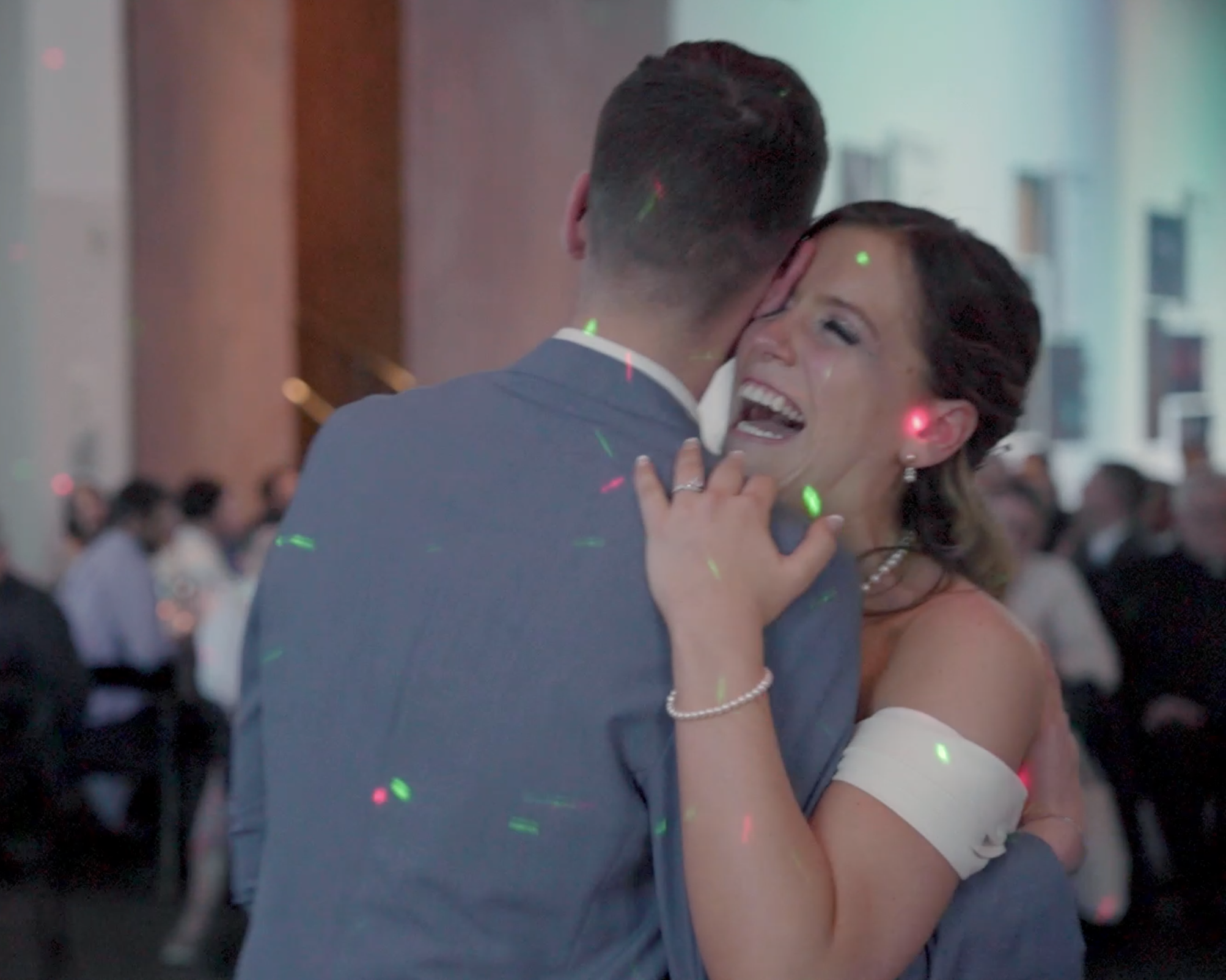 A woman and man dance closely at a wedding reception, with the woman smiling and laughing while the man has his back to the camera, in a decorated hall with other guests seated in the background.