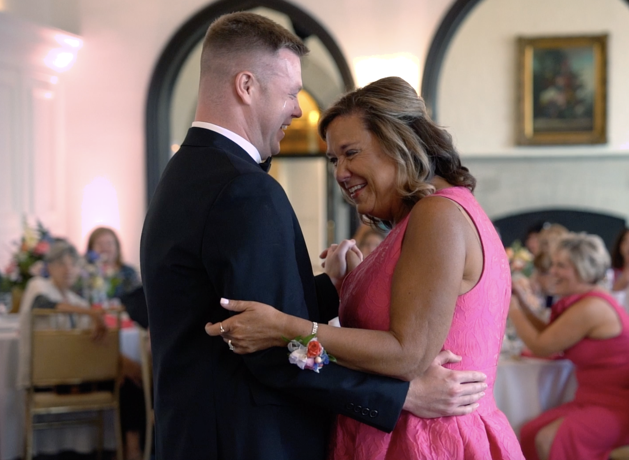 A young man in a black tuxedo dancing with an older woman in a pink dress at a wedding reception, both smiling and holding hands.