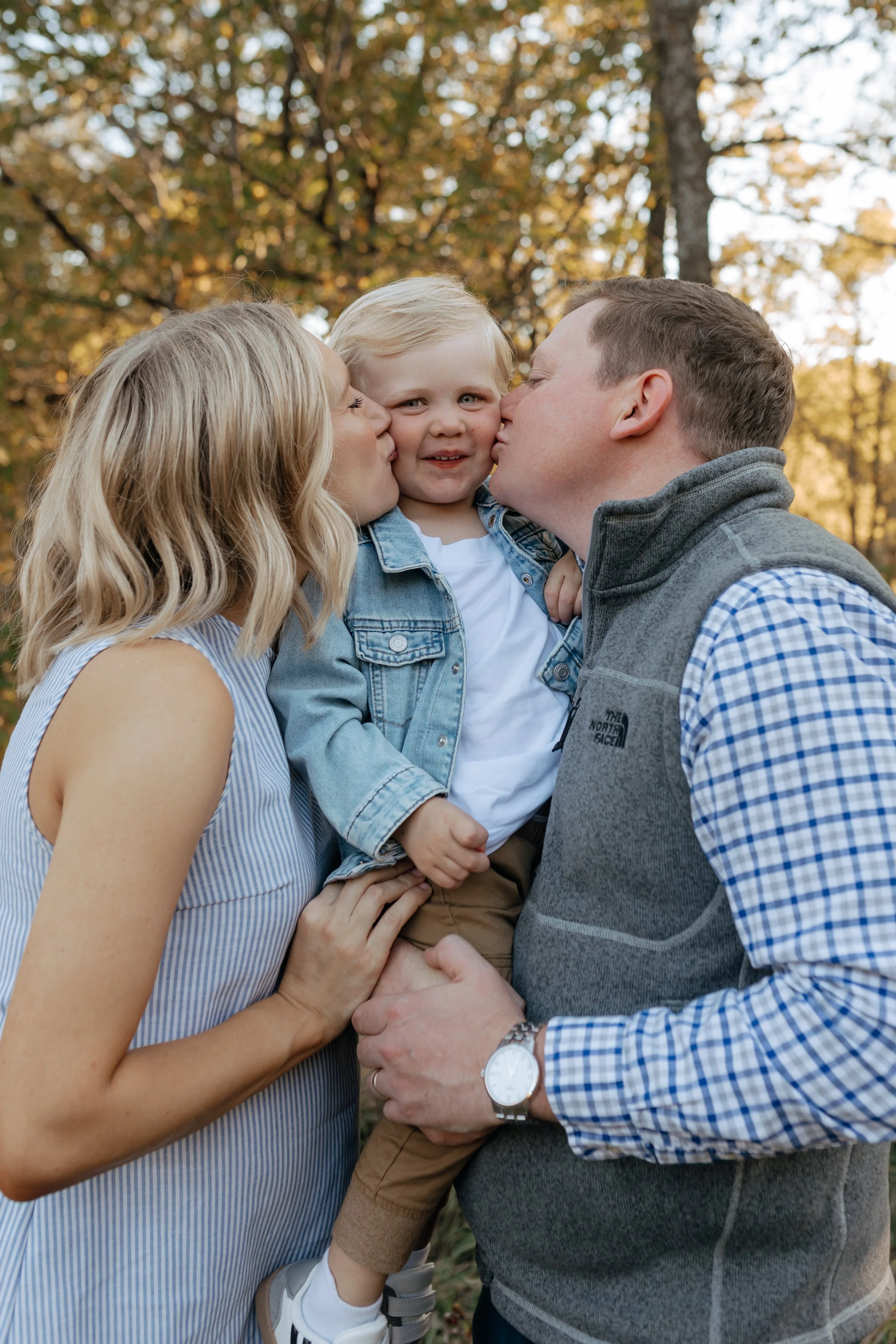A family of three, consisting of a woman, man, and young child, sharing a kiss outdoors during fall, with trees with autumn leaves in the background.