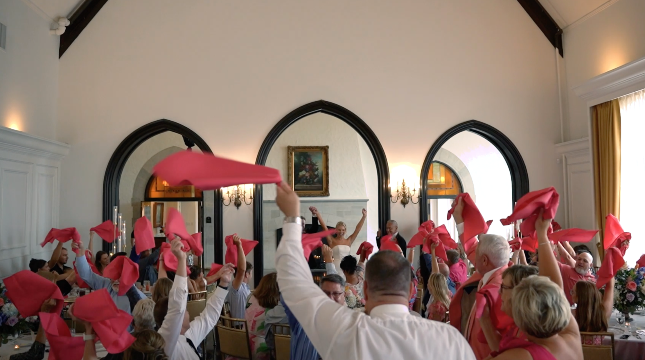 Wedding celebration with guests waving pink napkins in a decorated banquet hall.