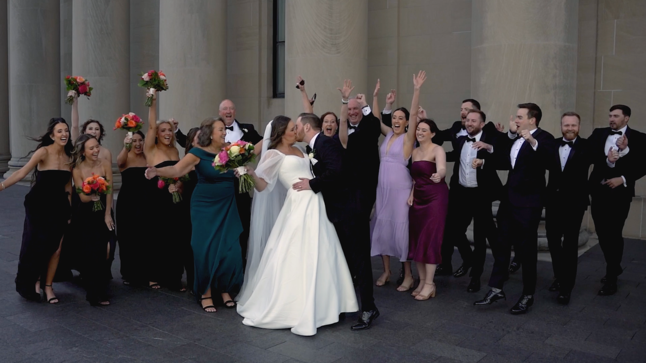 A wedding celebration with a bride and groom kissing surrounded by celebrating friends and family. The group is outdoors against a stone building, with some holding bouquets and raising their hands in excitement.