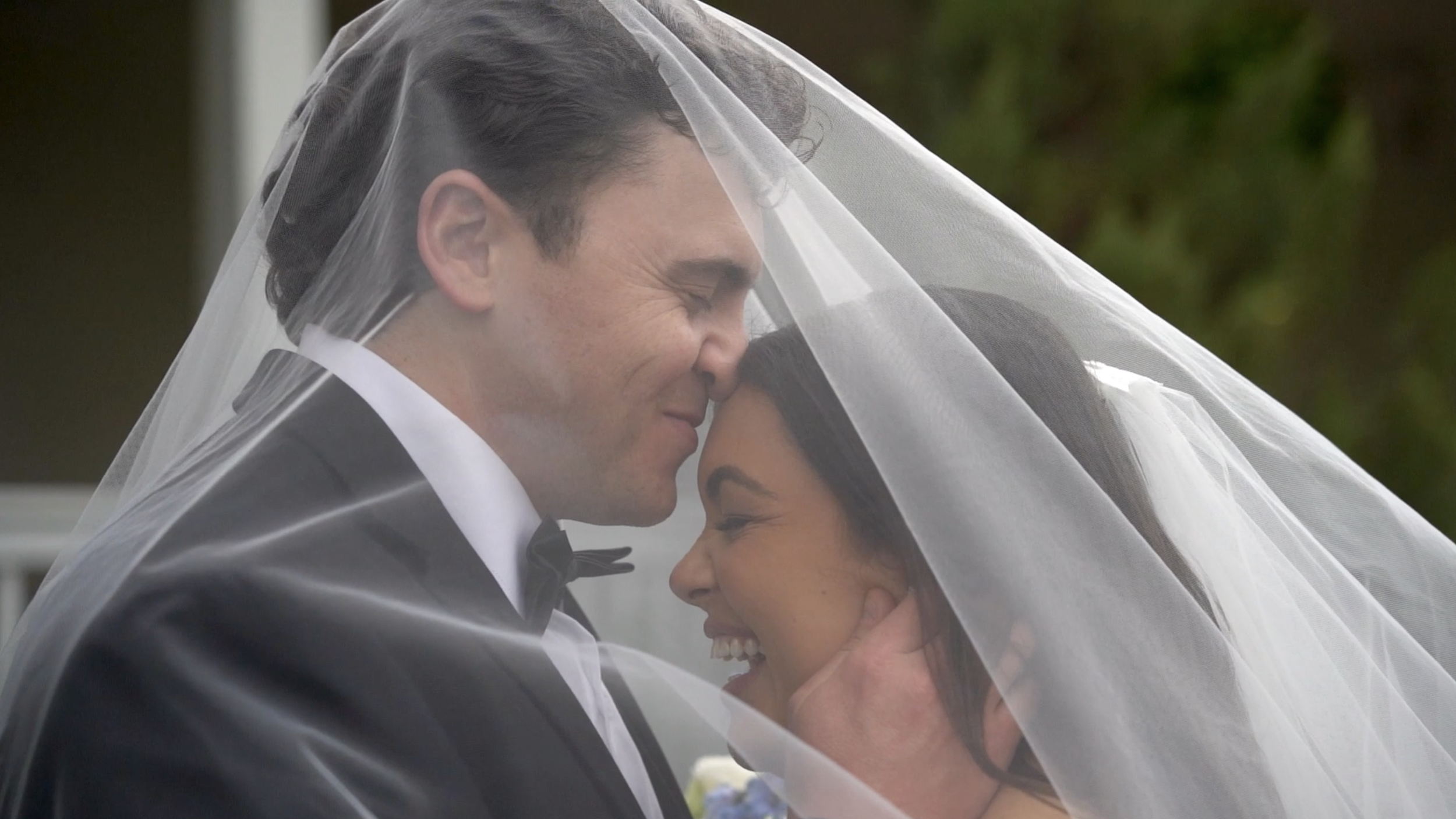 A newlywed couple embracing underneath a white wedding veil, smiling and touching foreheads, outdoors with greenery in the background.