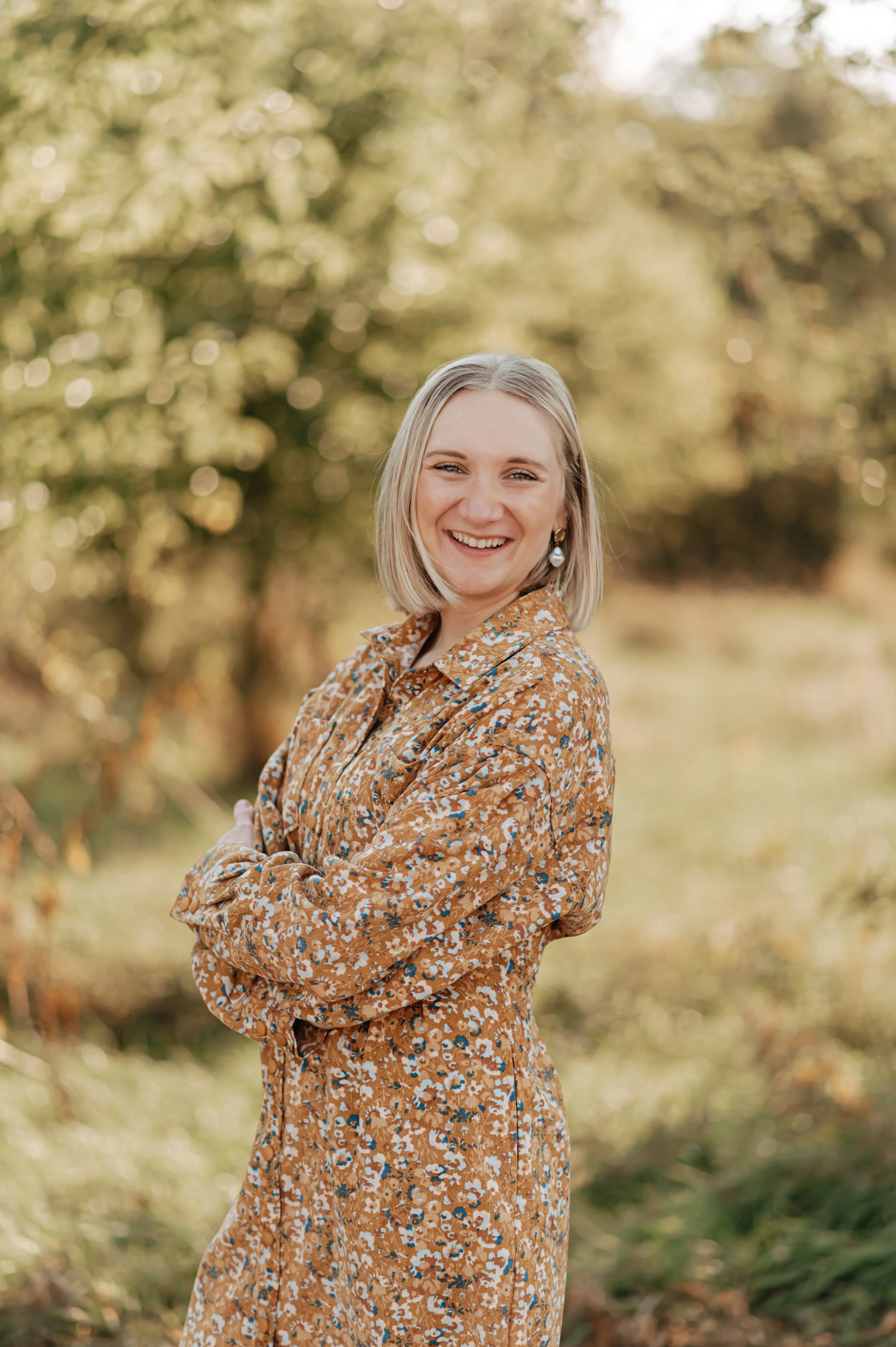 A woman with blonde hair smiling, standing outdoors in a natural setting with blurred trees and foliage in the background, wearing a brown floral dress.