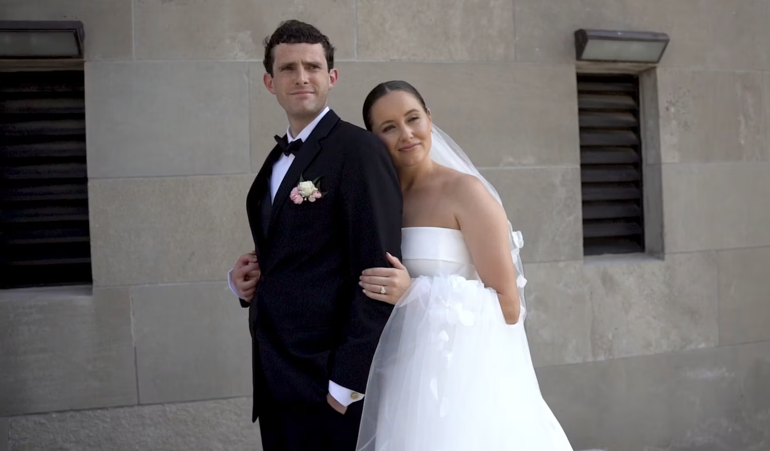 A newlywed couple standing against a stone wall, with the bride leaning on the groom's shoulder and both smiling softly.