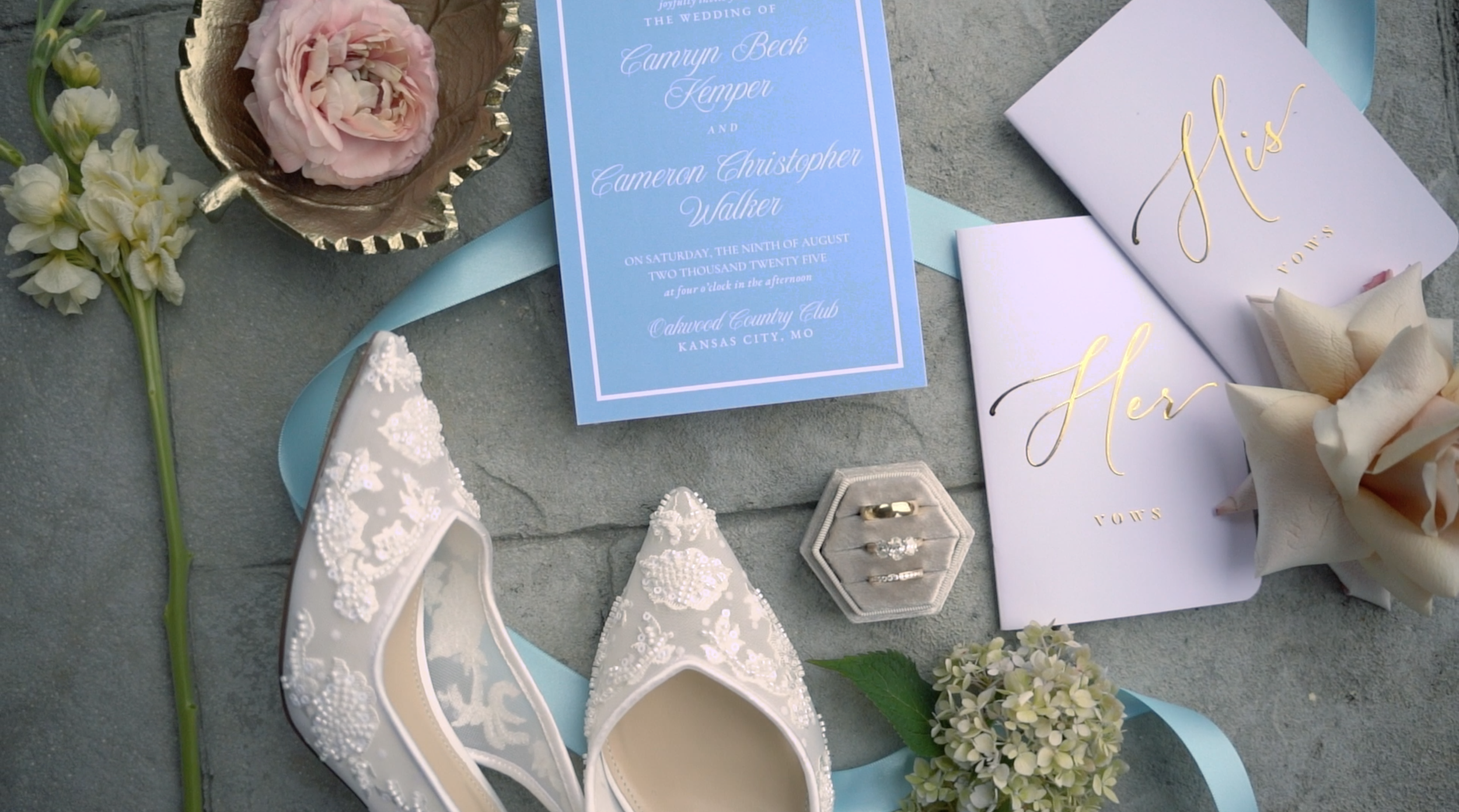 Wedding invitation cards, a pair of embroidered wedding shoes, a ring box with rings, a bouquet, a small flower arrangement, and a medal, arranged on a gray surface with a light blue ribbon.