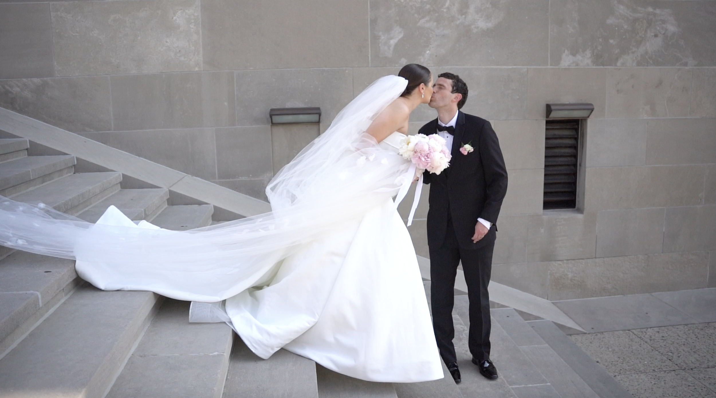 A bride and groom sharing a kiss on a set of stone stairs outside a building. The bride wears a white wedding dress with a long train and veil, holding a bouquet of pink and white flowers. The groom is dressed in a black tuxedo with a bow tie and boutonniere.
