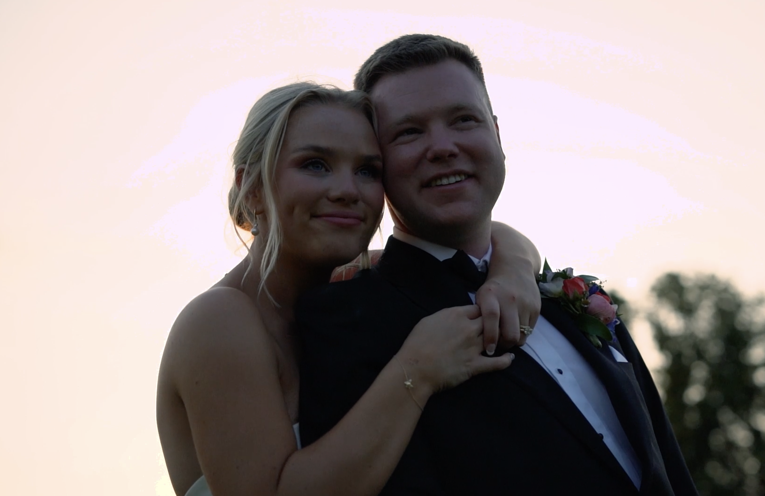 A smiling bride and groom on their wedding day, embracing outdoors at sunset with trees in the background.