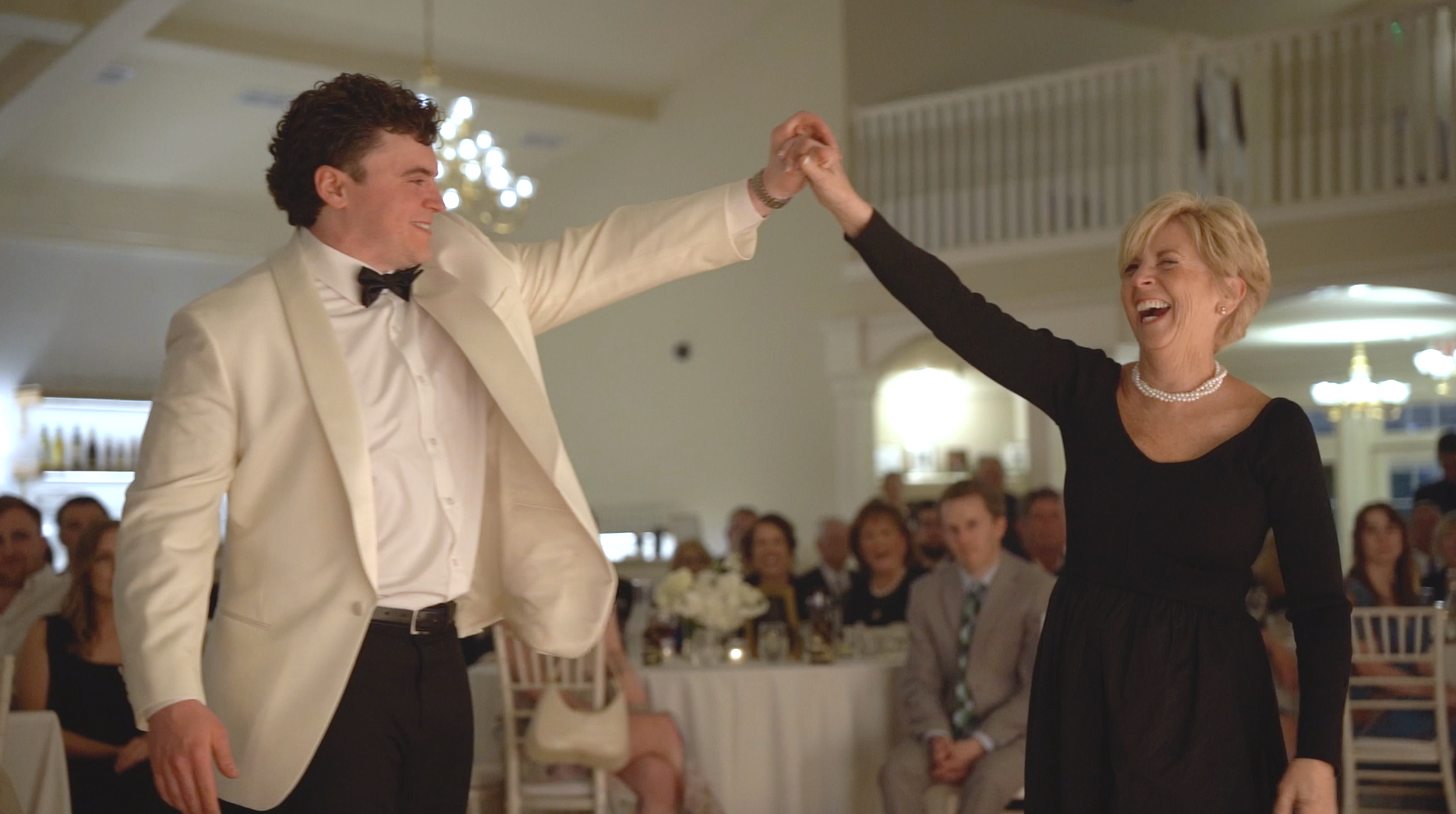 A man in a white tuxedo and a woman in a black dress dancing together while holding hands and smiling, with an audience in the background.
