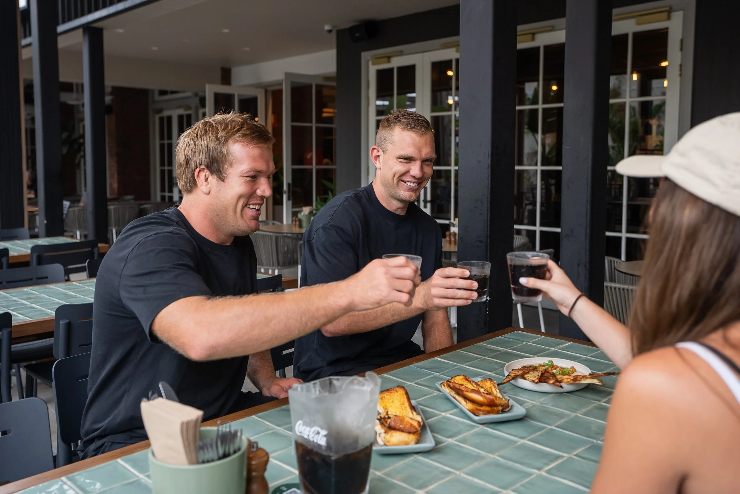 Three men in black shirts sitting at a table outdoors, smiling and raising glasses filled with dark soda, toasting each other, with a woman in a white hat whose face is not visible, with plates of food including sandwiches and pizza on the table.