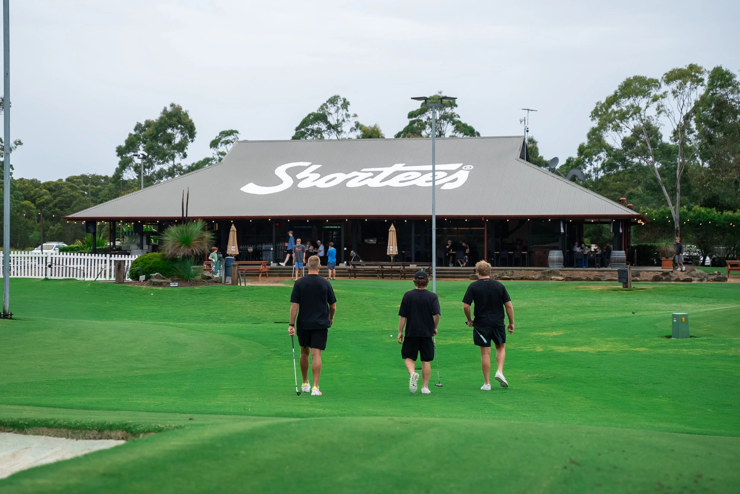 Golf course with four people walking on the green, practice putt, in front of a building with a large 'Stubb' logo on the roof, which appears to be a restaurant or clubhouse, surrounded by trees.