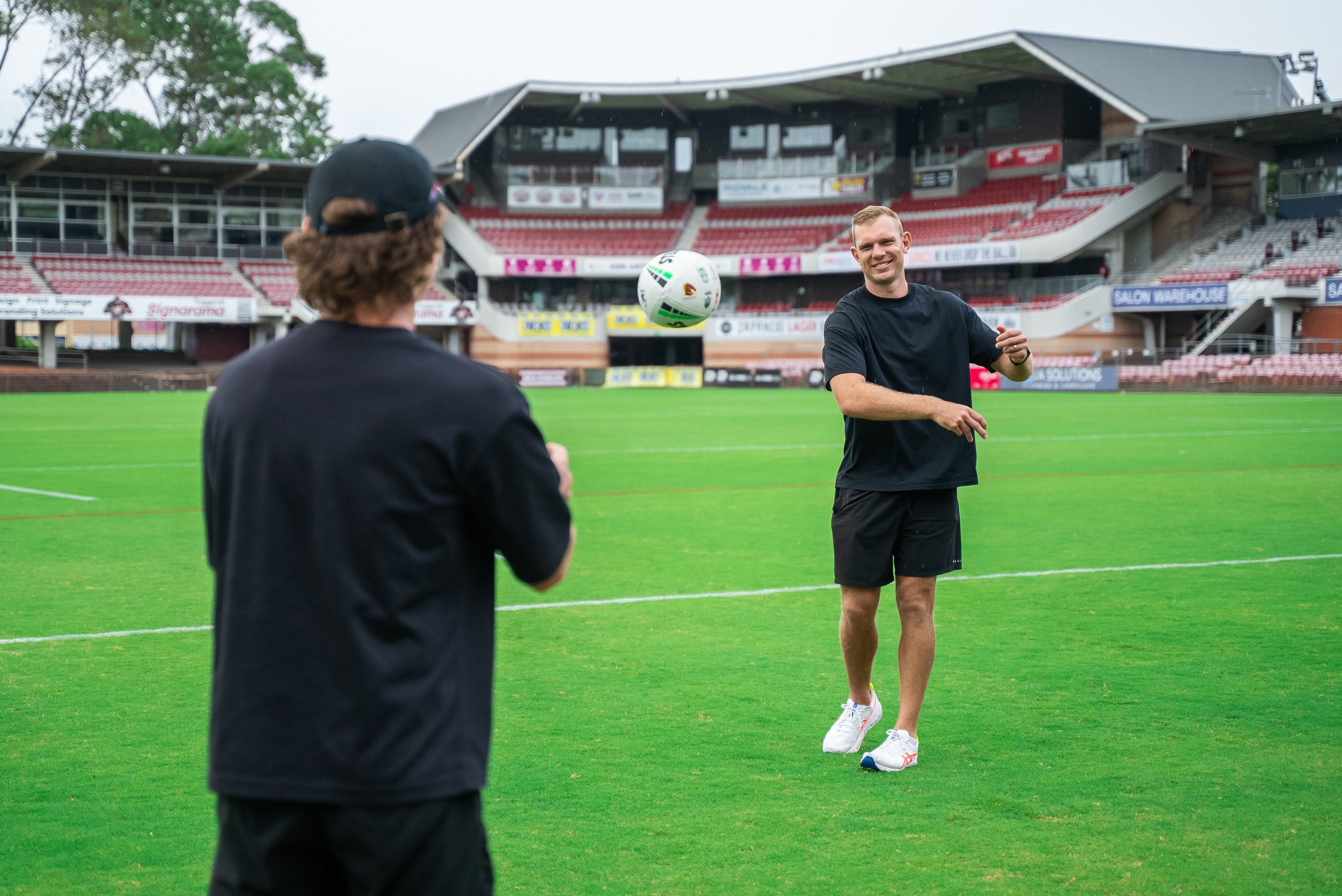 Tom Trbojevic smiling and standing on a sports field, engaging in a game involving a ball, with another person in the foreground whose back is to the camera. The setting is a stadium with empty red seats and advertising banners.
