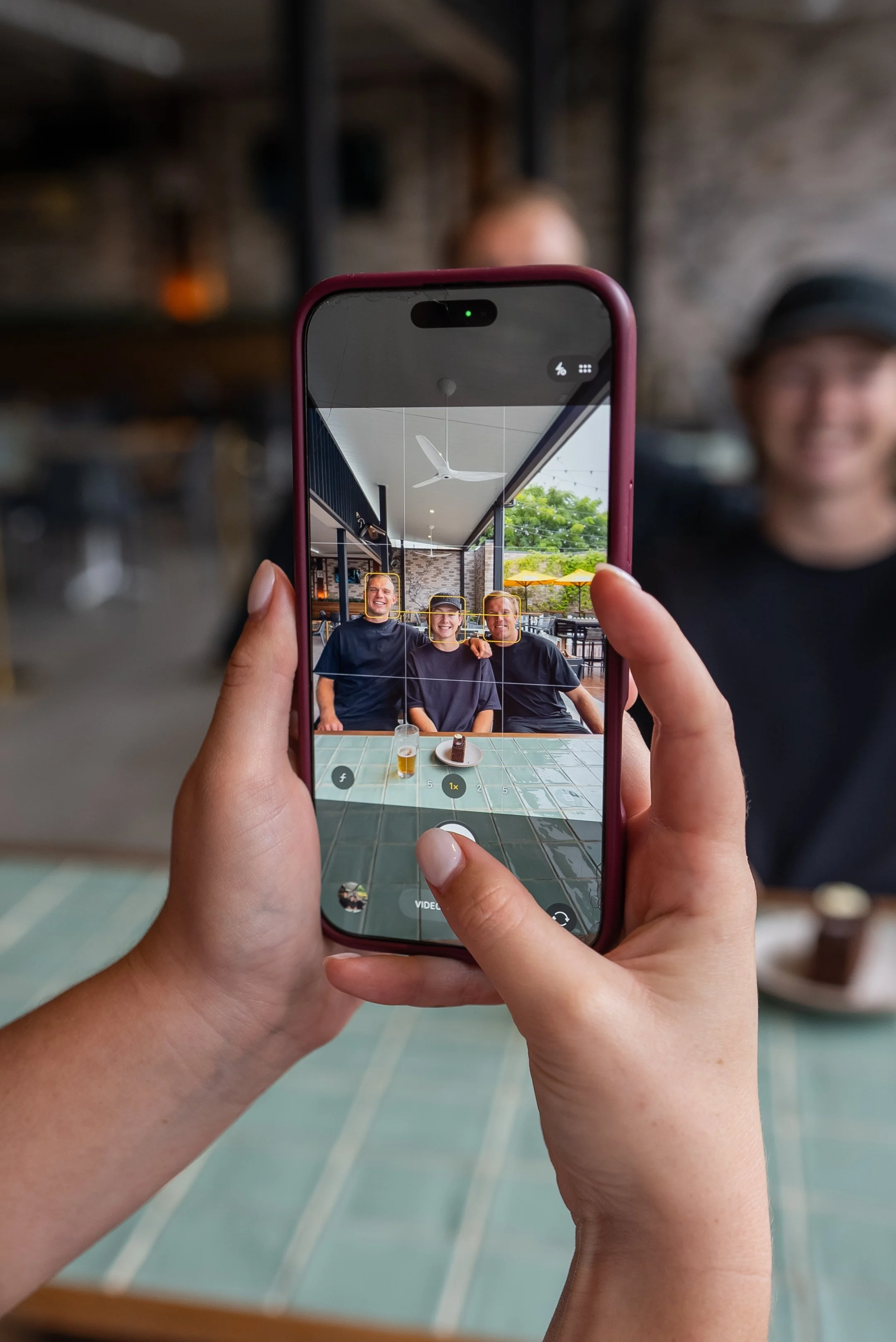 A person taking a photo of three friends sitting at a table inside a restaurant or cafe. The camera screen shows the friends smiling and sitting close together.