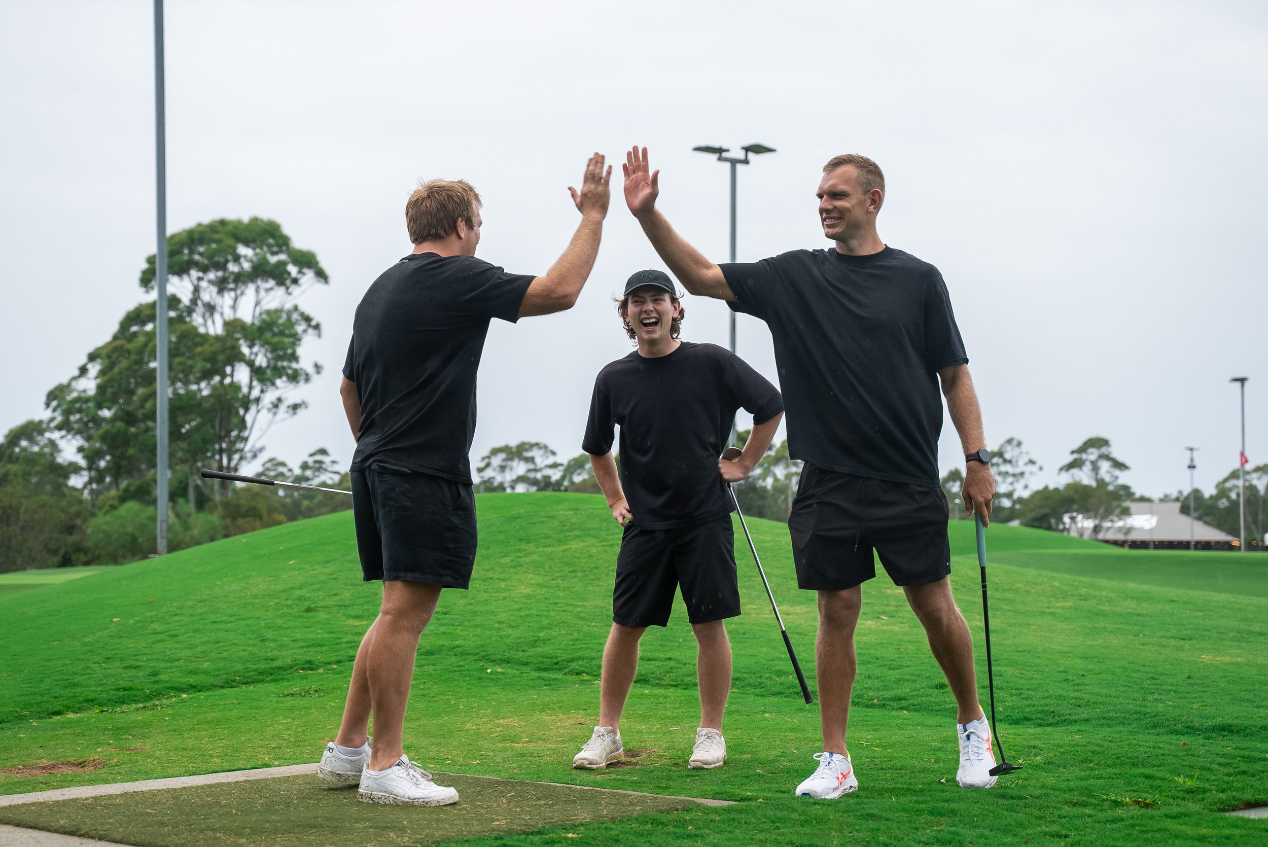 Three young men on a golf course, two giving each other a high five, one smiling with a golf club, all wearing black t-shirts and athletic shoes, overcast sky.