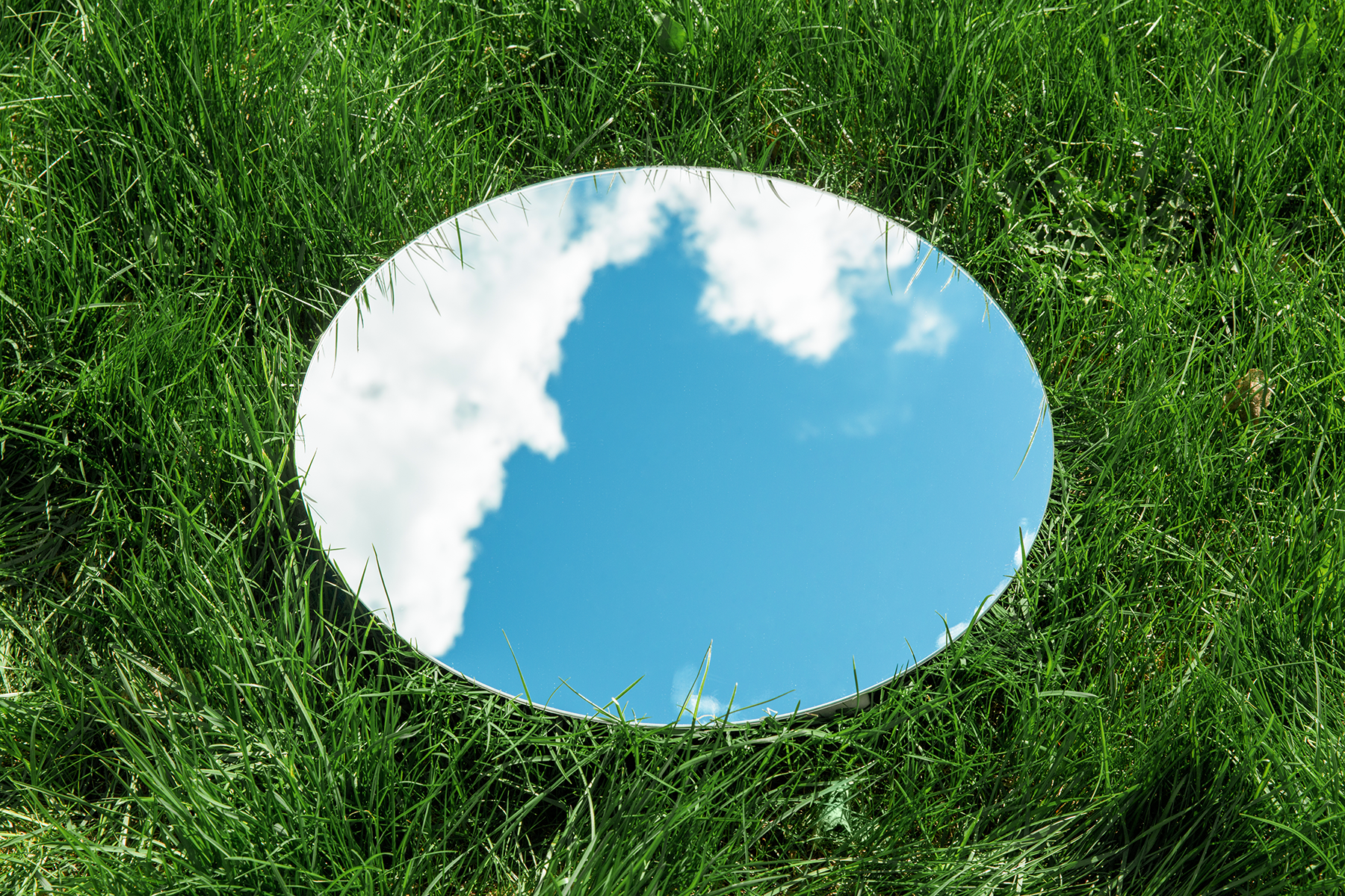 A round mirror lying on green grass, reflecting a blue sky with white clouds.