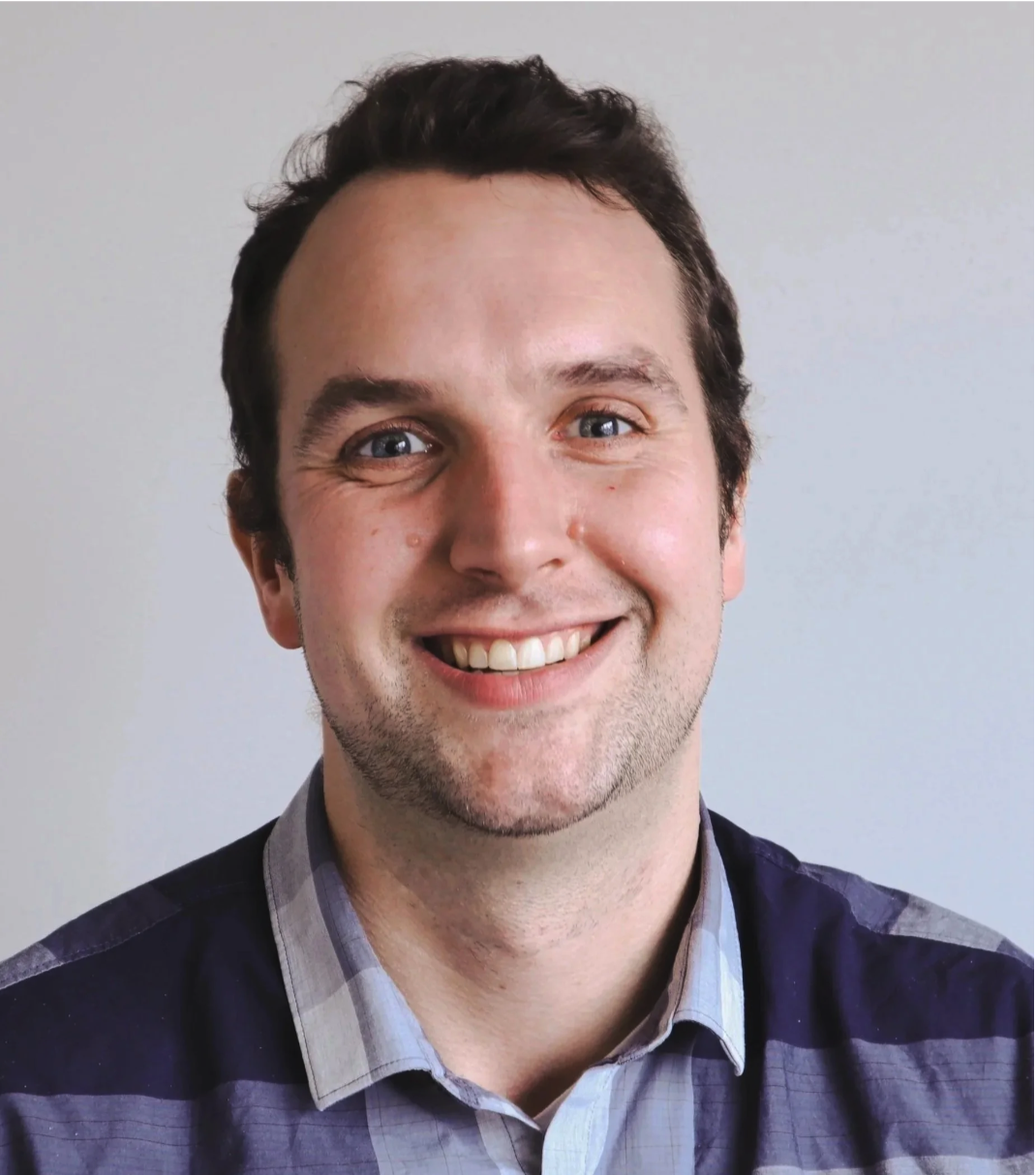 A close-up of a smiling man with dark hair, blue eyes, and light skin, wearing a plaid shirt, against a plain light background.
