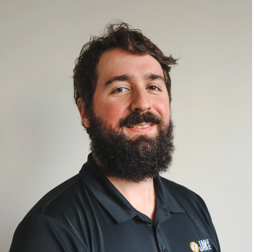 A man with dark hair, a full beard, wearing a black polo shirt with a logo, smiling and looking at the camera, standing in front of a plain light-colored wall.