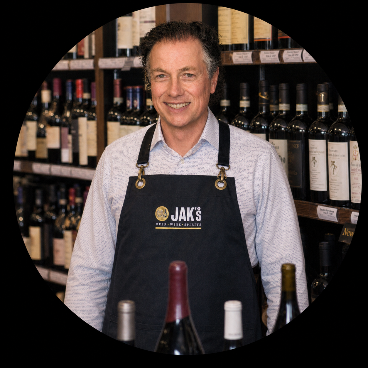 A man smiling in a wine and spirits store, wearing a black apron with 'JAK's Beer Wine Spirits' logo.