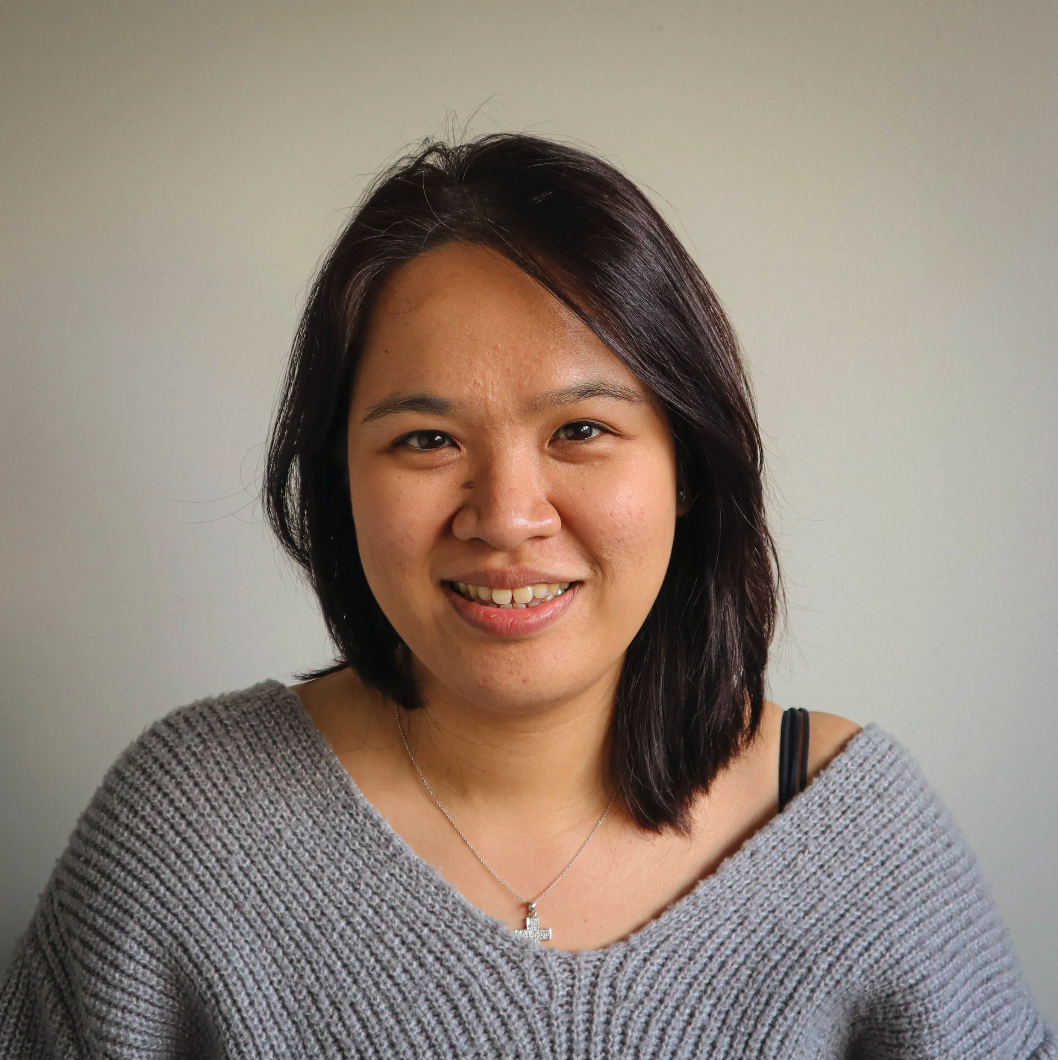 A woman with shoulder-length dark hair smiling at the camera, wearing a gray sweater and a cross necklace, standing against a plain light background.