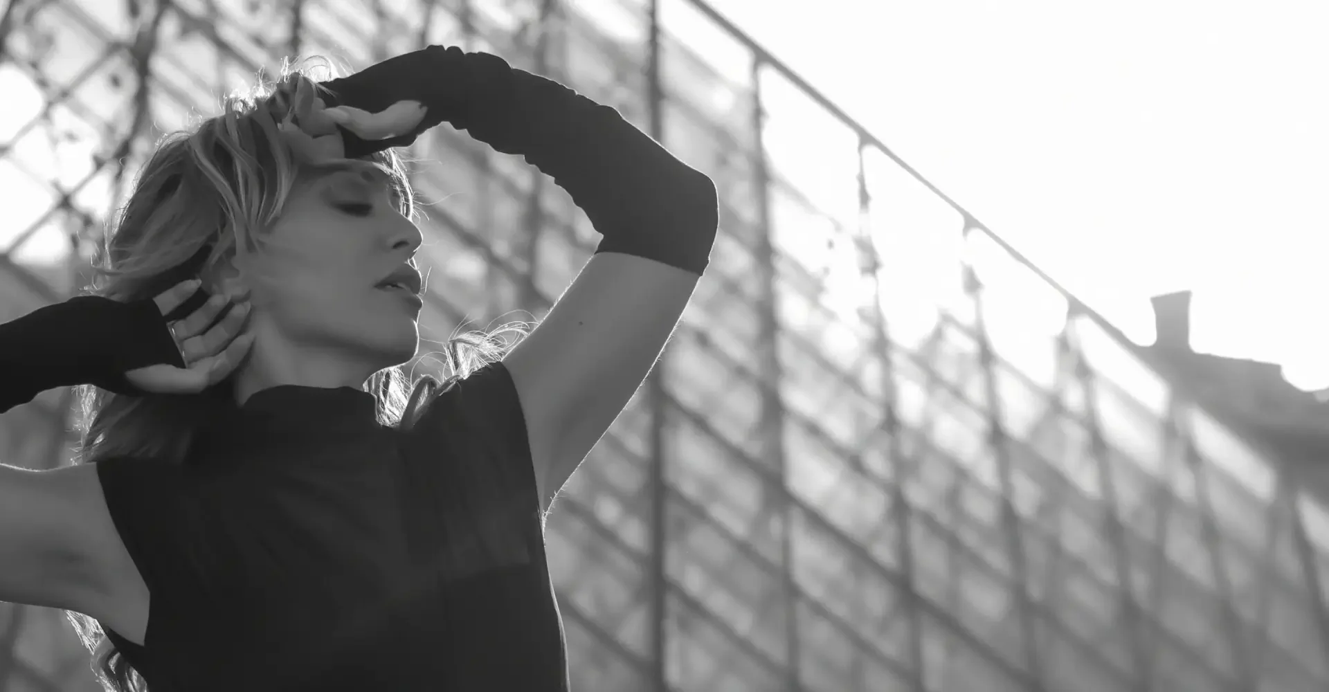 Black and white photo of Rakel Chafir  with long hair, wearing a short-sleeved top and gloves, posing with her eyes closed and one hand on her forehead, against a background of a building with a glass roof.