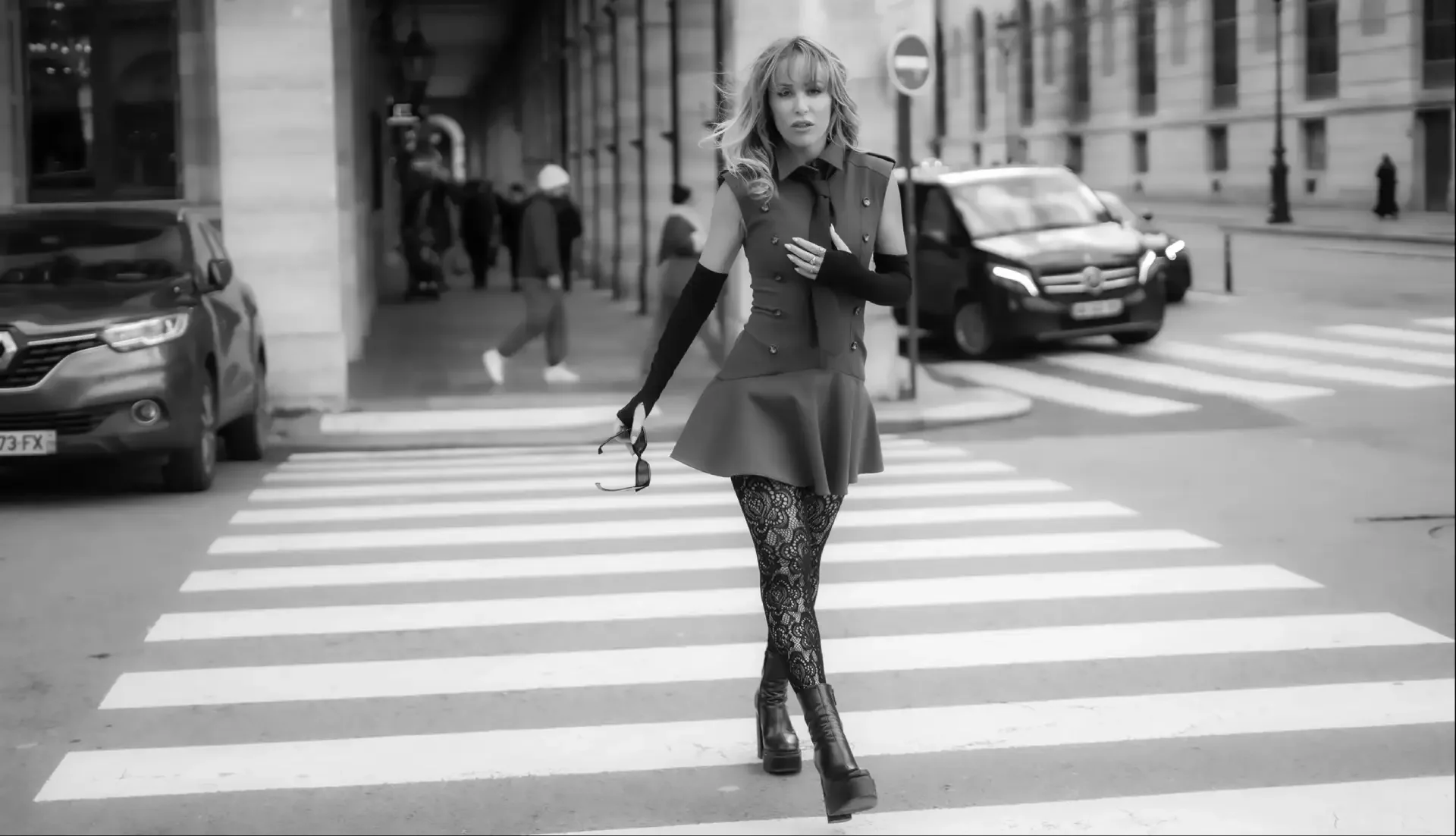 Black and white photo of Rakel Chafir  crossing the street on a zebra crossing in an urban setting, holding sunglasses and wearing leather boots, a dress, and long gloves.