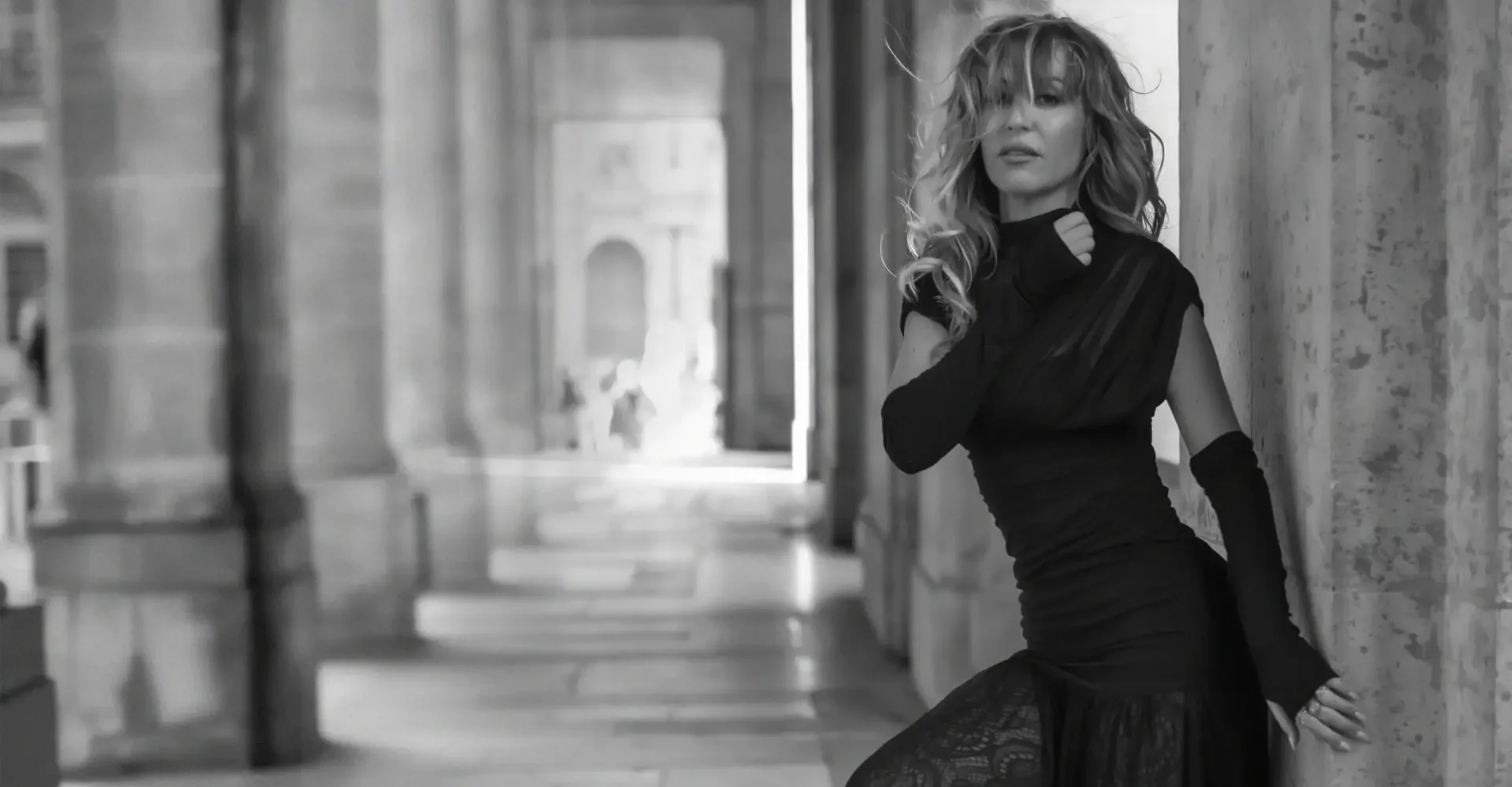 Rakel Chafir  with wavy blonde hair leaning against a stone pillar in a black dress with lace details and long black gloves, with an urban architectural background in black and white.