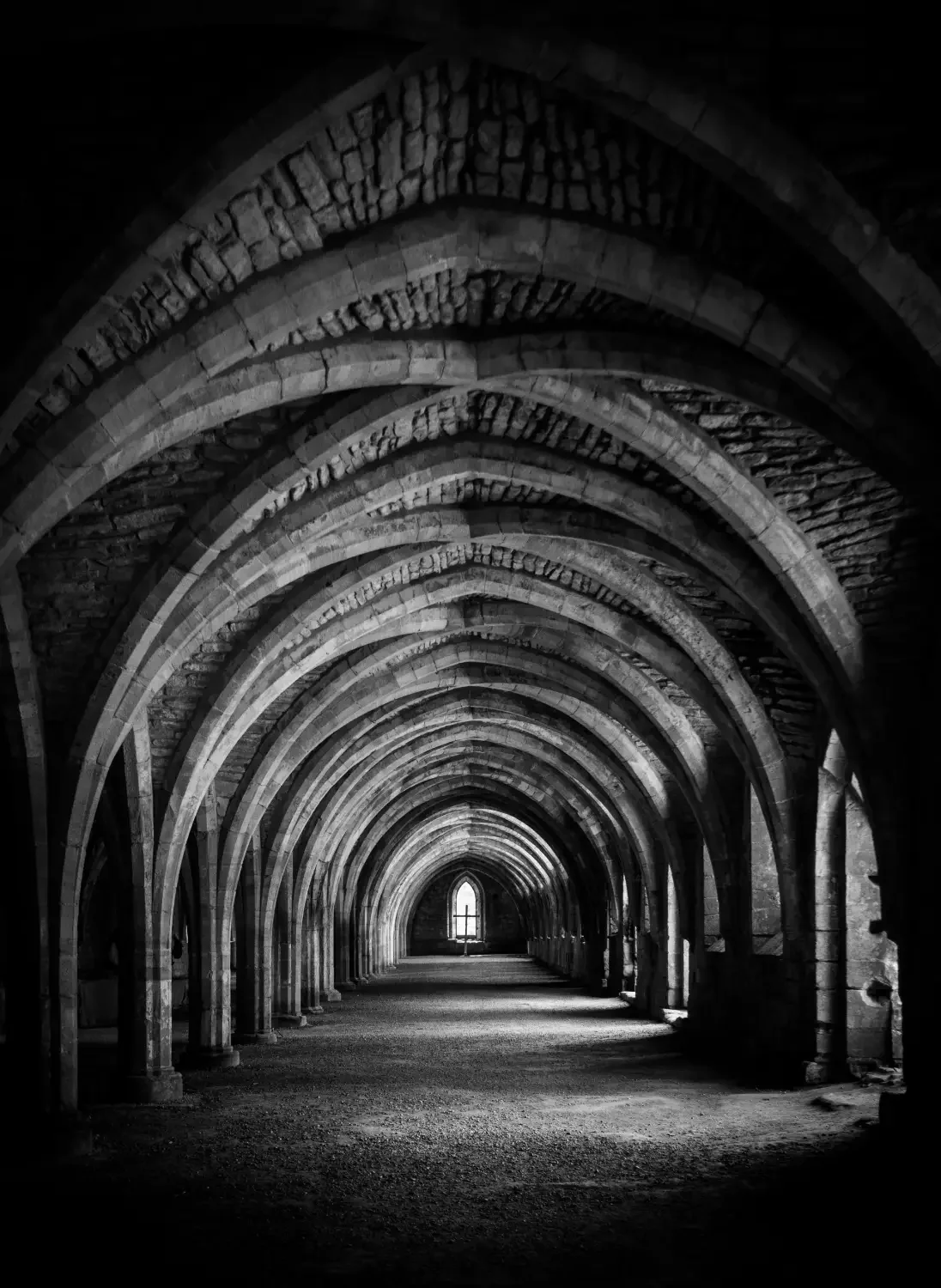 A black and white photo of a Gothic-style arched hallway with stone columns and pointed arches, leading to a small window at the end.