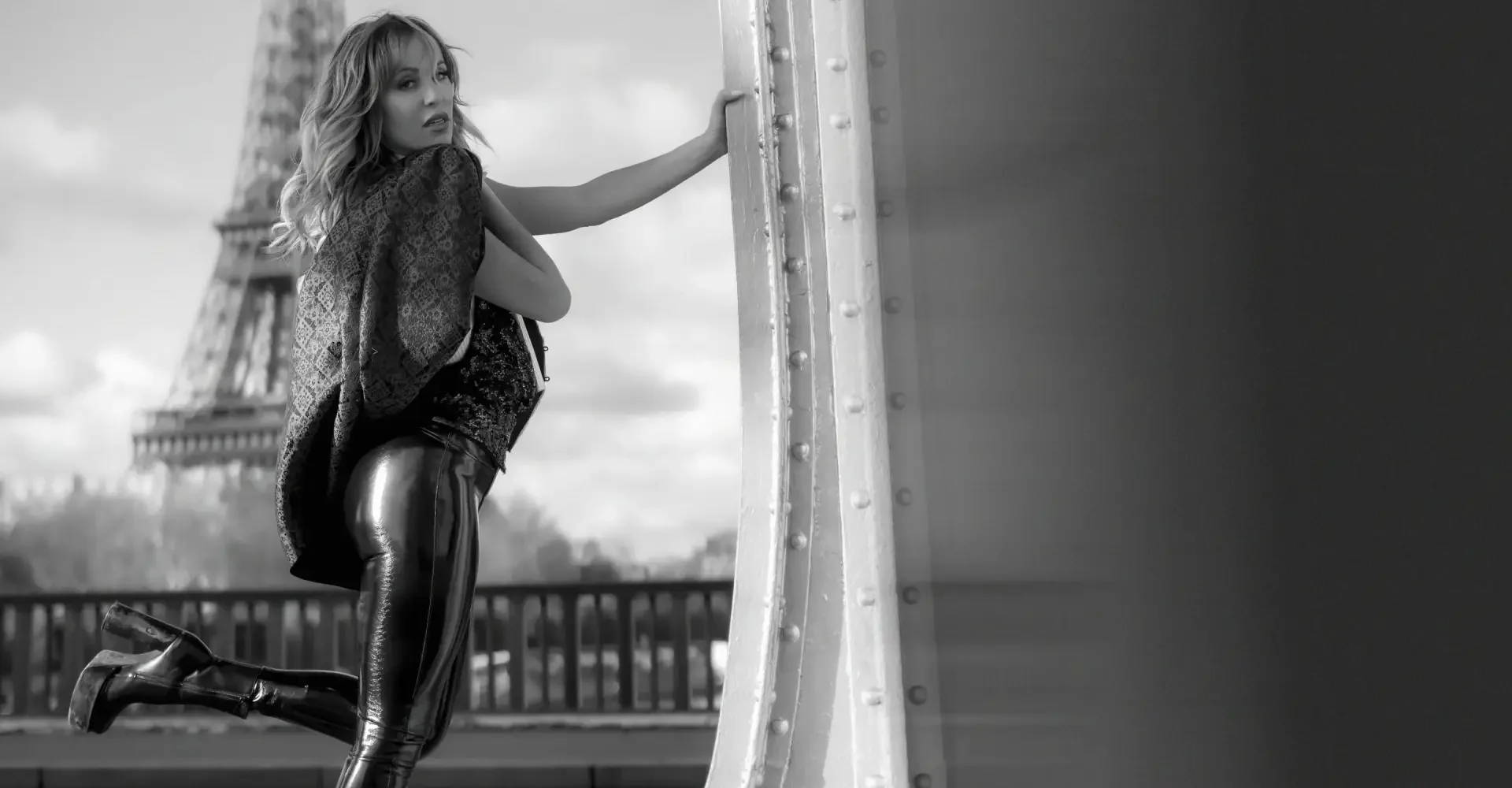 A woman wearing shiny black leggings, a patterned jacket, and boots kneeling on one knee on a bridge near the Eiffel Tower in Paris, France, with a hand on a metal pillar.